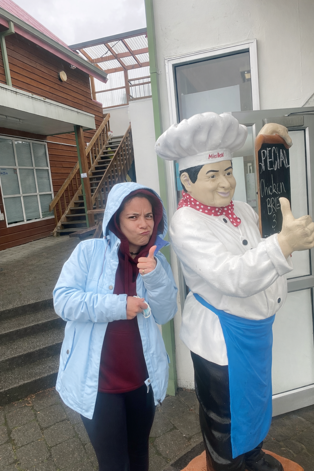 A woman in a blue jacket and hood, making a skeptical face and pointing at the camera, standing next to a large chef statue outside a restaurant, with a menu board that reads "Special Chicken Brea".