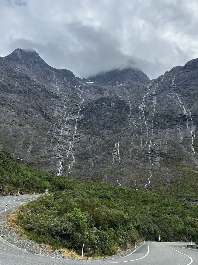 A winding mountain road with lush green bushes in the foreground, a steep rocky mountain with multiple small waterfalls, and overcast sky above.
