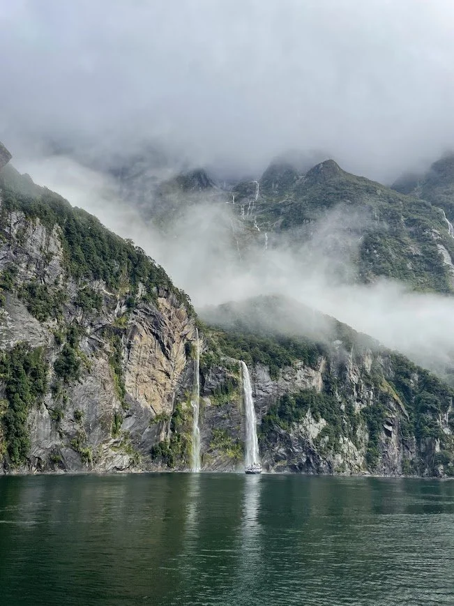 A misty mountain scene with waterfalls, steep cliffs, and calm water in the foreground.