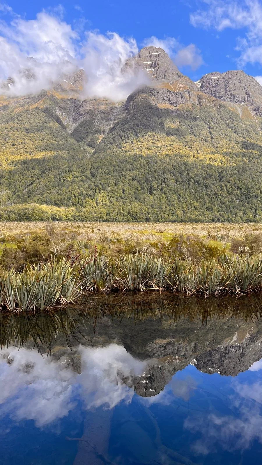 A mountain landscape with steep rocky peaks partly covered in snow, blue sky with some clouds, a forested hillside, and a body of water reflecting the scene.
