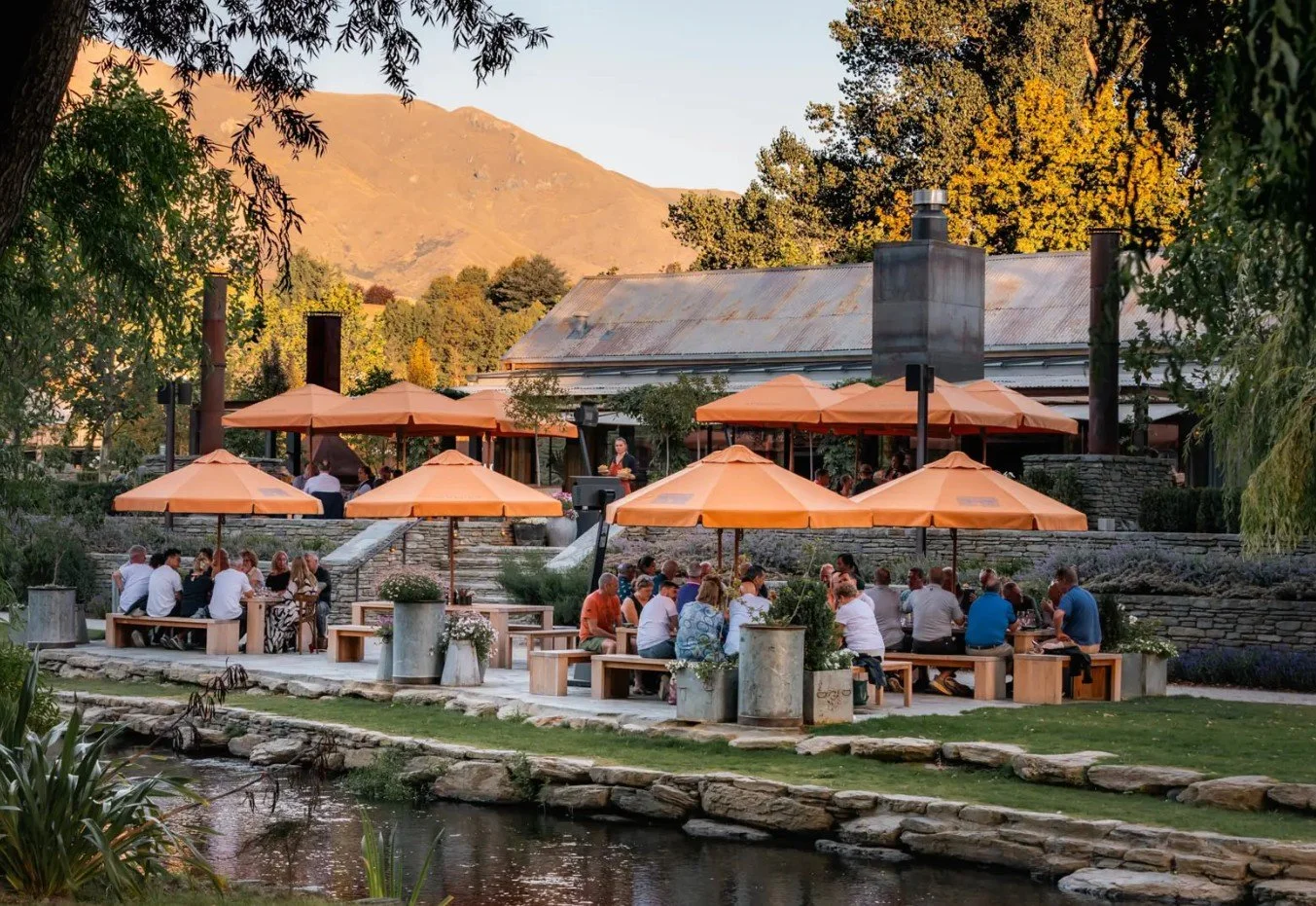 People dining outdoors at a restaurant with orange umbrellas, set along a riverbank, against a backdrop of trees and mountains.