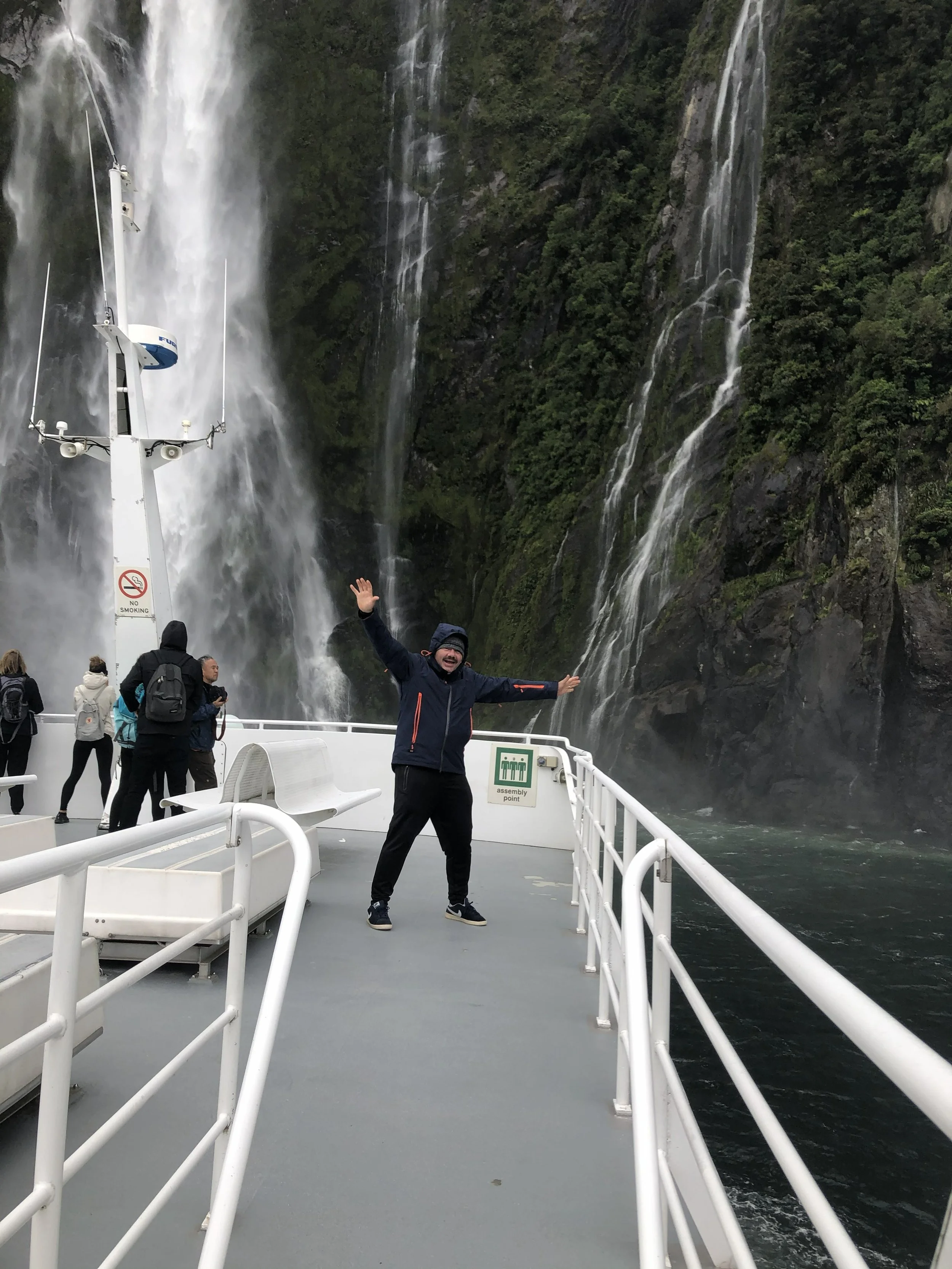 Person standing on a boat deck with arms raised, tourists behind, crashing waterfalls in the background.