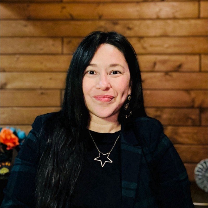 A woman with long black hair and light skin, smiling, wearing a black top, star-shaped necklace, and earrings, seated indoors against a wooden wall.