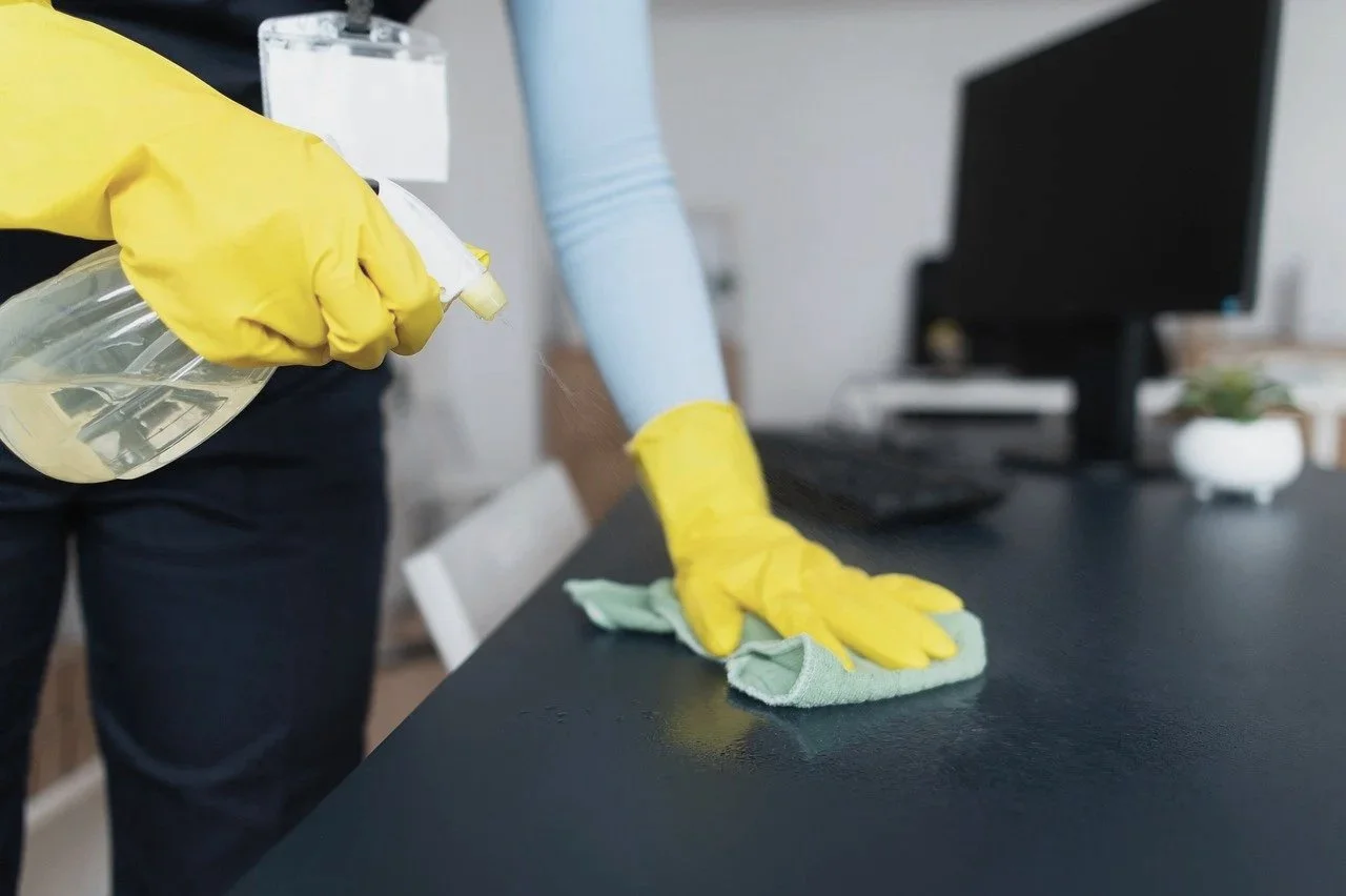 Henkilö puhdistaa mustaa pöytää keltaisissa suojahanskoissa ja vasemmalla kädellä pyyhkii pyyhkeellä, samalla kun oikeassa kädessä on sumutin.