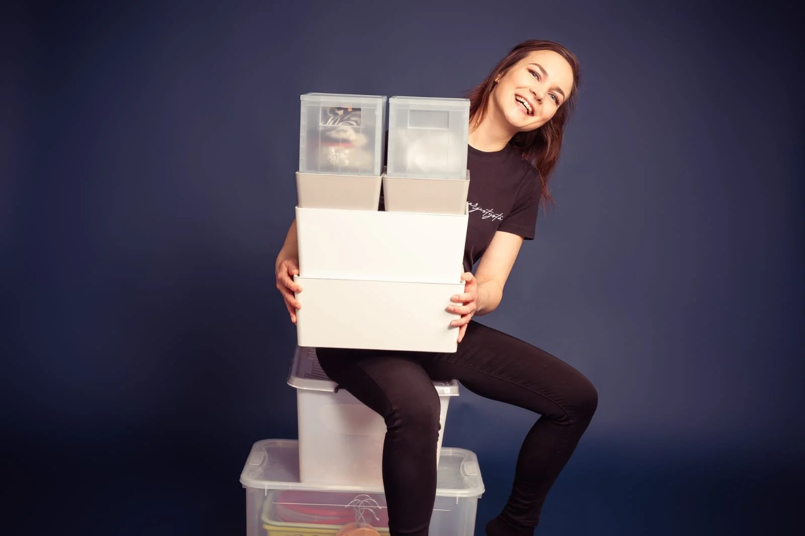 Iloinen nainen istuu laatikoiden päällä, pitäen samalla laatikoita sylissään ja hän hymyilee. Taustalla on tummansininen seinä.