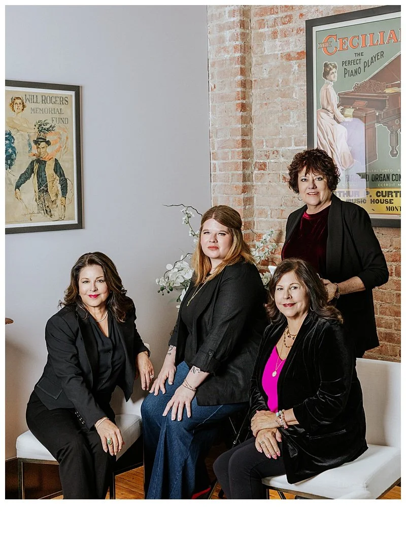 Four women in black and pink attire posing in a room with framed vintage posters, a brick wall, and white chairs.