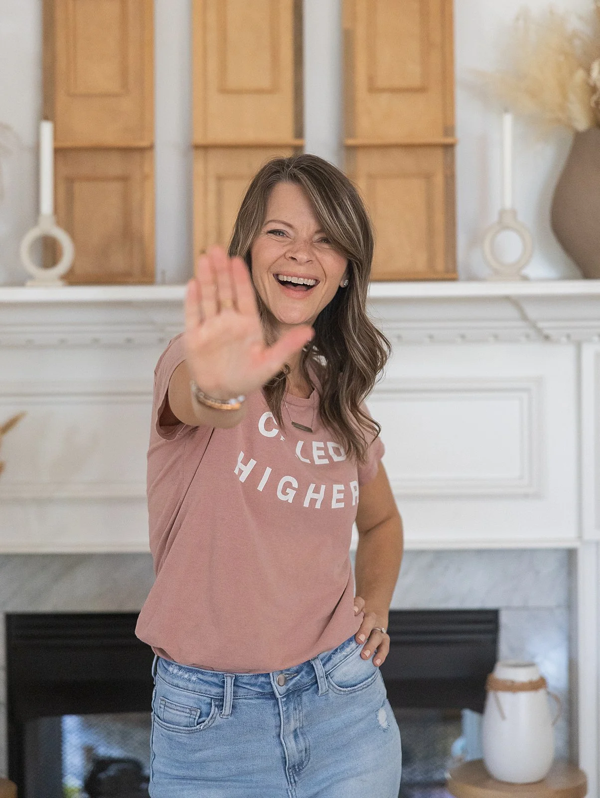 A woman with brown hair smiling and raising her hand towards the camera, wearing a pink t-shirt and blue jeans, standing in front of a fireplace with a white mantel and wooden cabinets in the background.