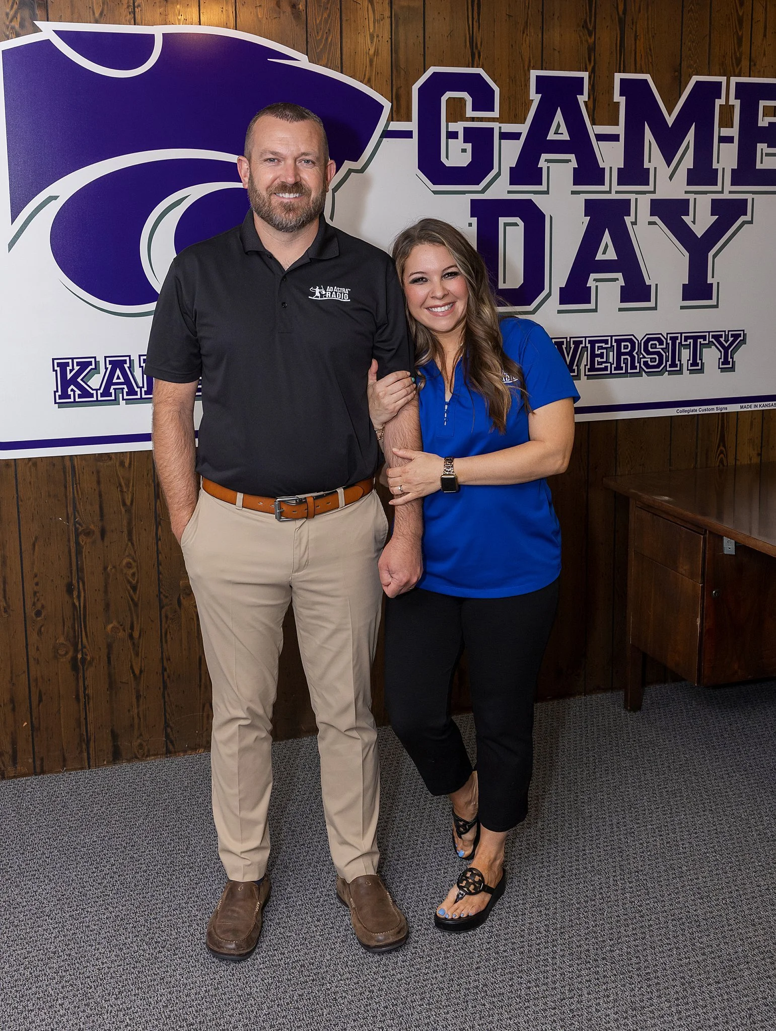 A man and woman standing together inside a room with wooden walls and a Kansas State University sign in the background. The man is wearing a black polo shirt and khaki pants, and the woman is wearing a blue polo shirt and black pants. The woman is holding the man's arm and both are smiling.