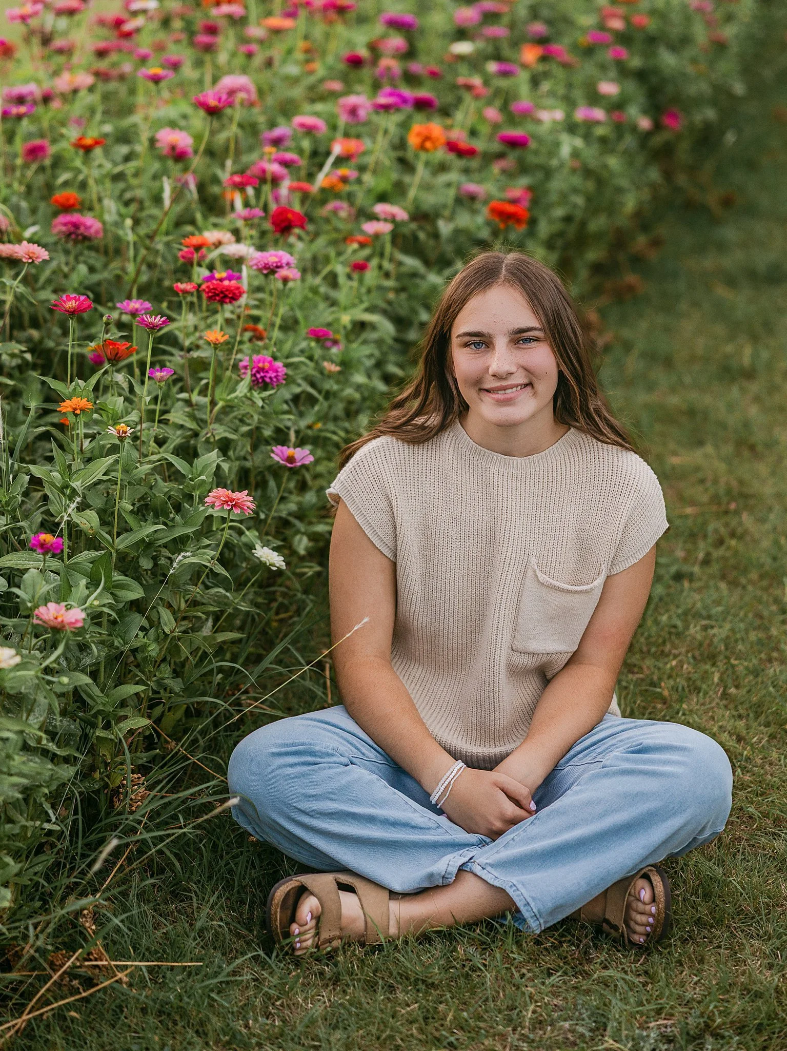 A young woman sitting cross-legged on the grass in front of a colorful flower garden, smiling at the camera.