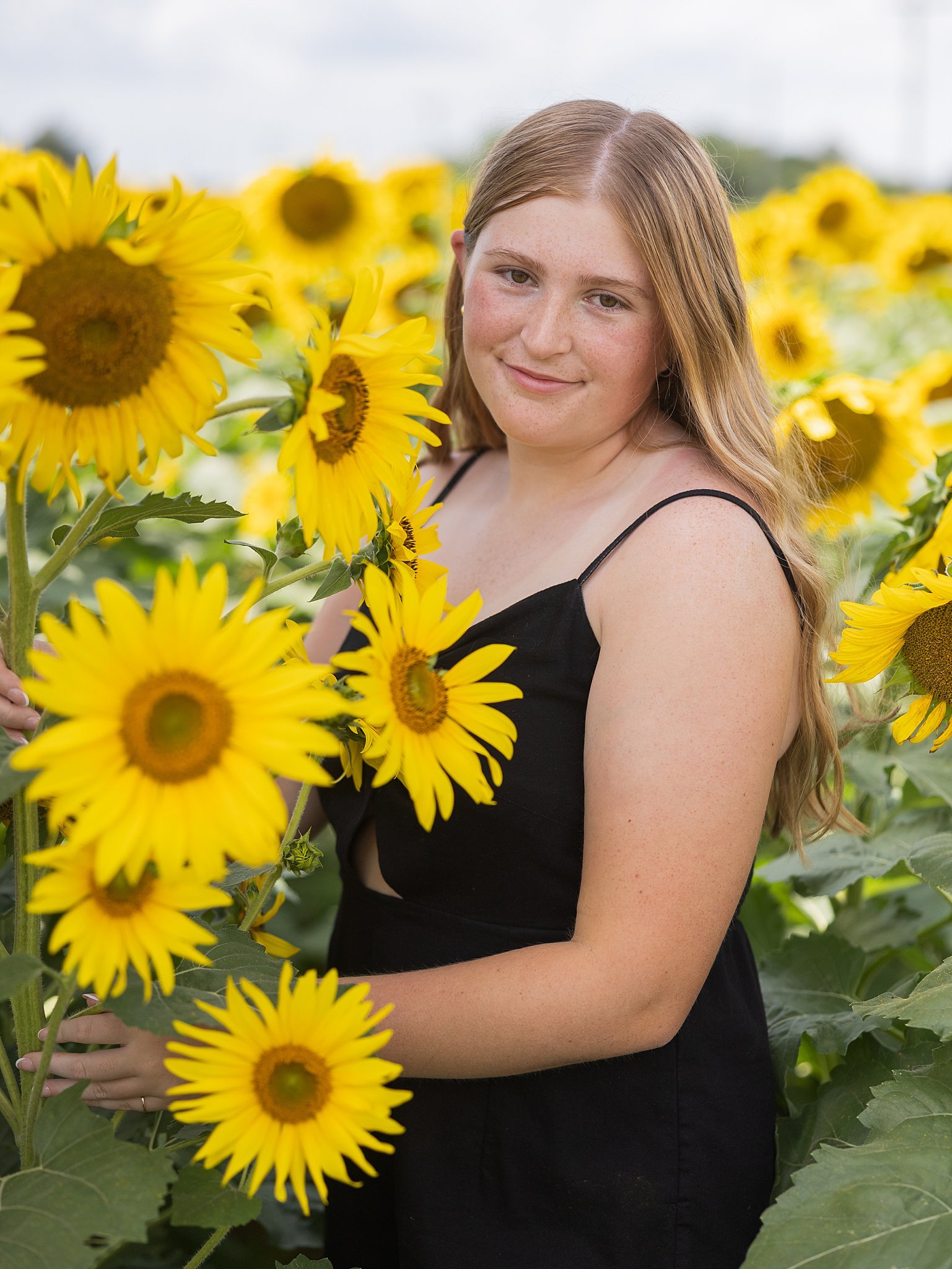 A young woman with long red hair and a black dress stands in a sunflower field, smiling at the camera.