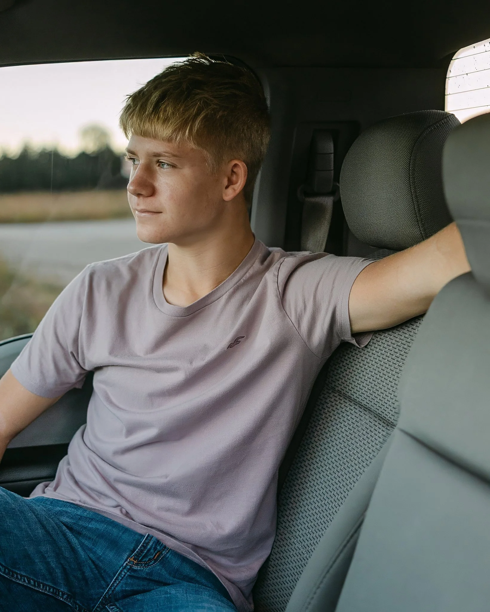 High school senior sitting in the back of a truck, looking out the window with a relaxed expression in natural light.