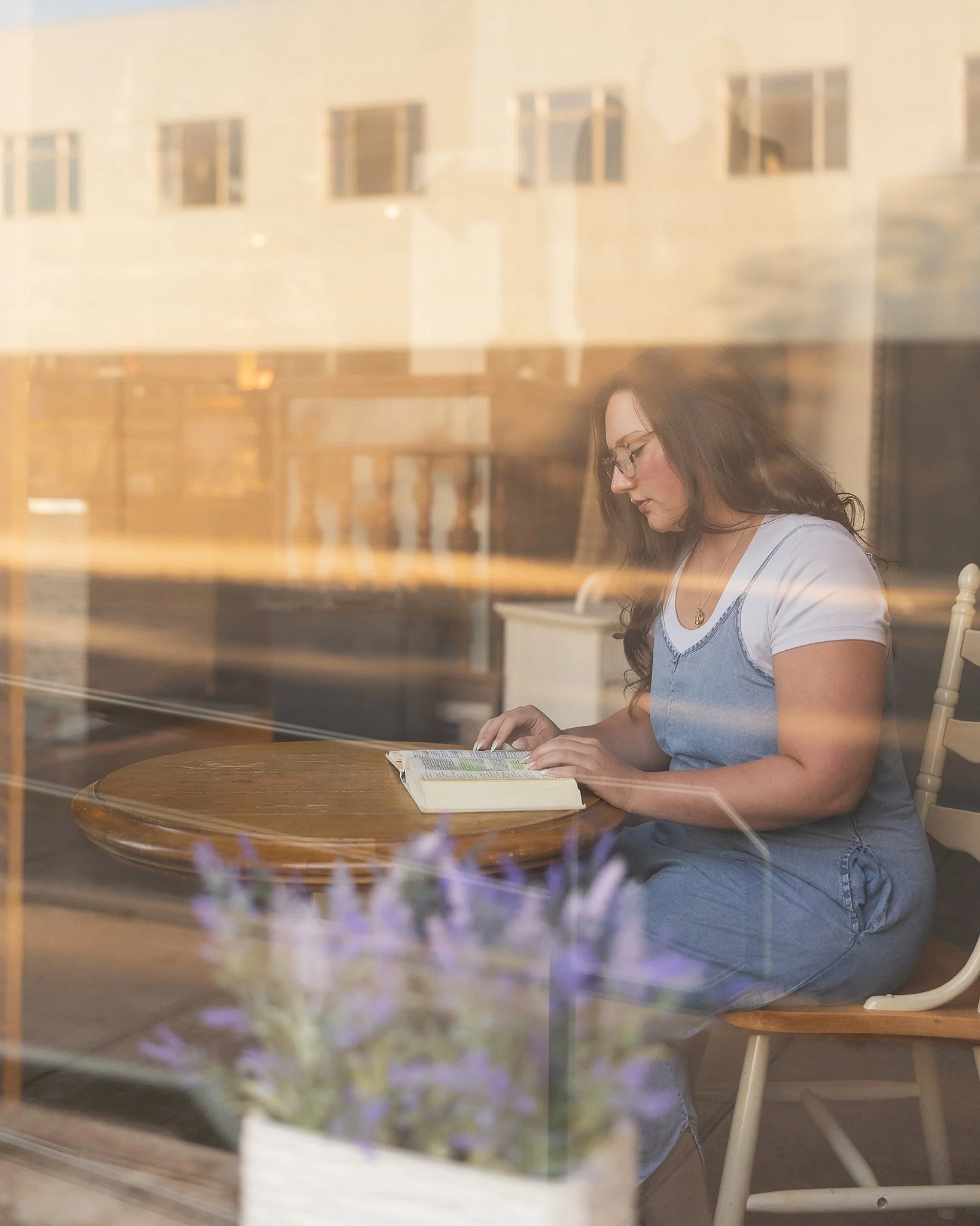 HIgh school senior girl 2026 with glasses reading a book at a table in a cozy cafe, seen through a window with reflections of buildings and a blurred purple flower in the foreground.