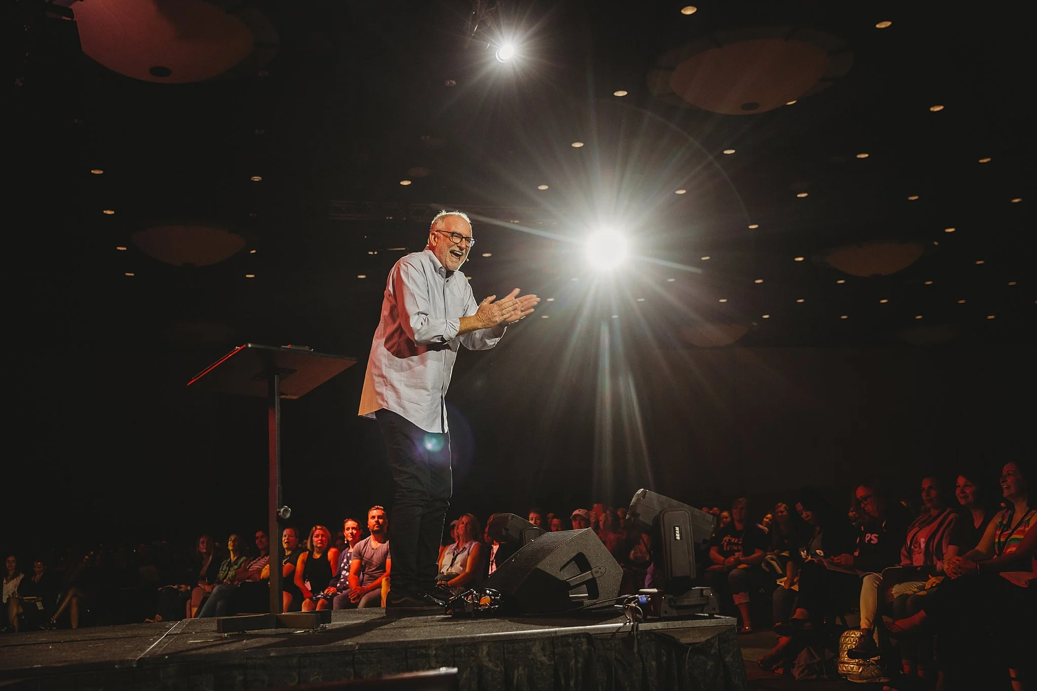 A man on stage clapping and laughing during a presentation or speech at a conference or event, with bright lights shining behind him and an audience of diverse people watching attentively.