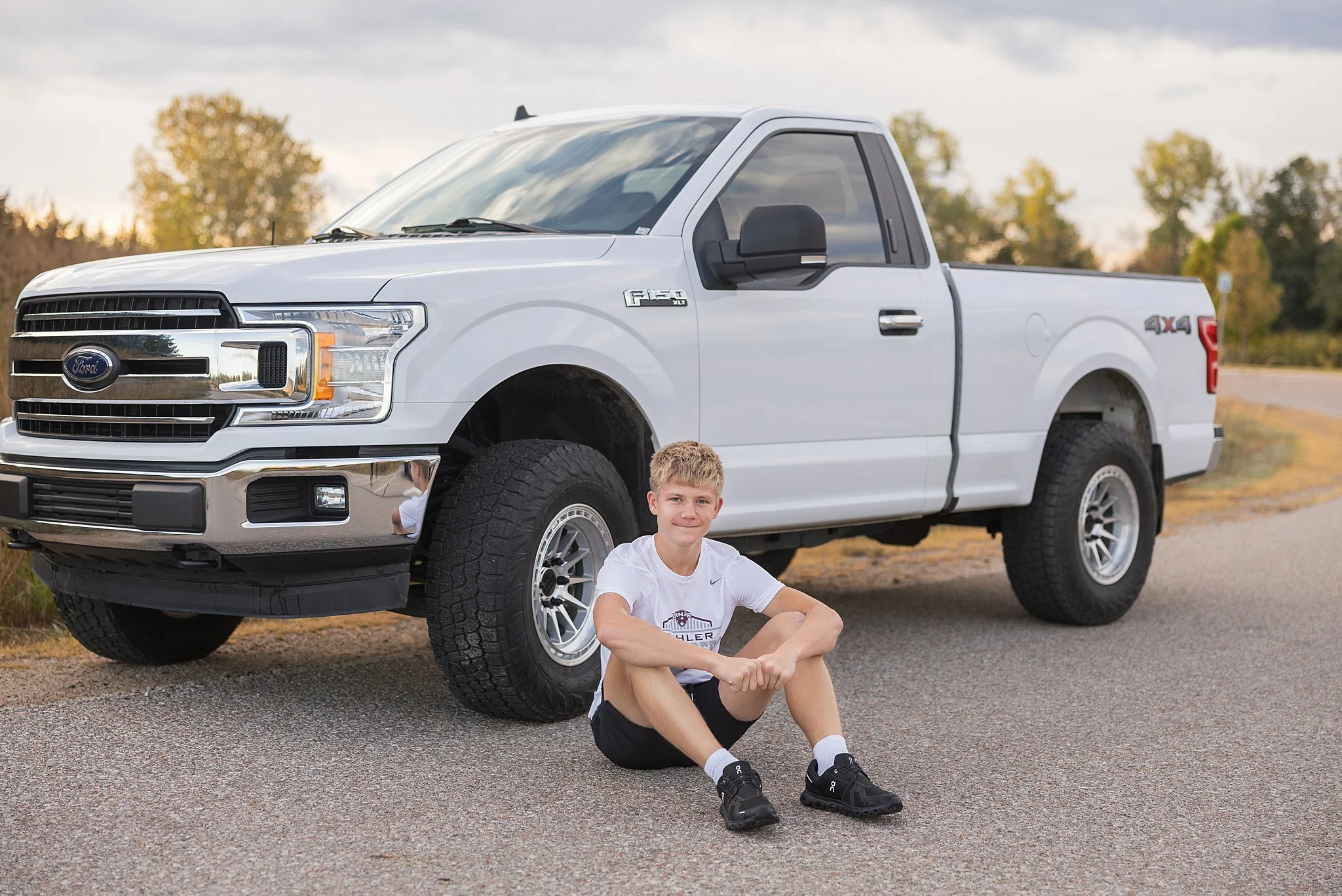 A young man sitting on the ground smiling in front of a white Ford F-150 pickup truck parked on the side of a rural road during daytime.