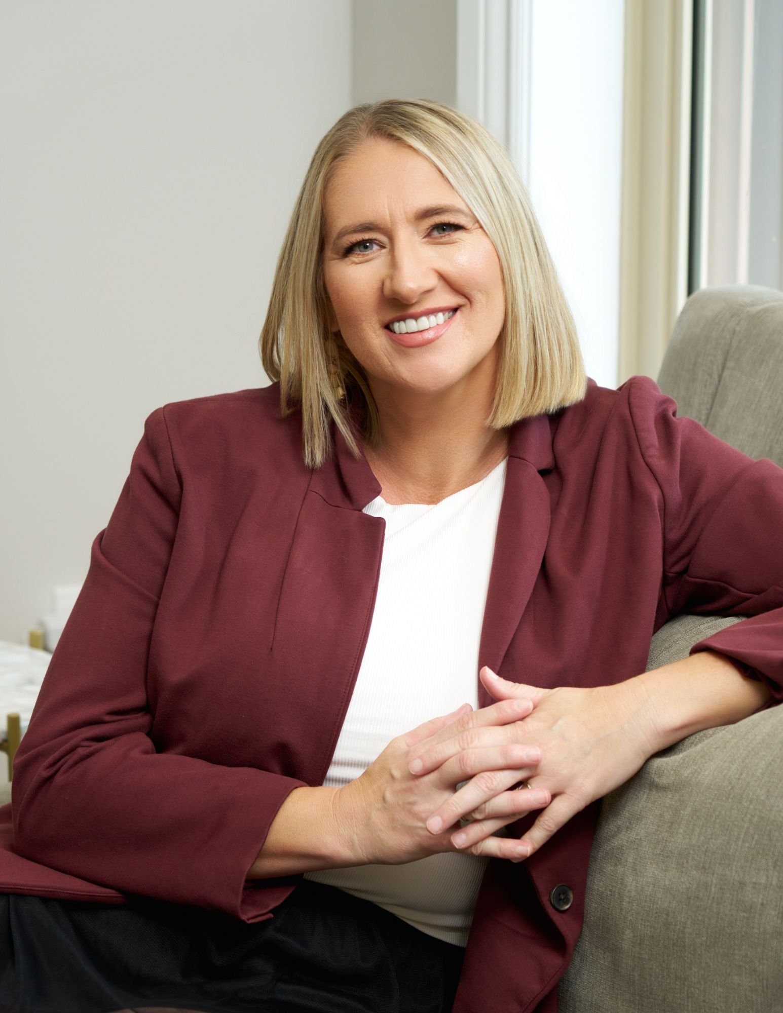 A smiling blonde woman wearing a burgundy blazer and white top, sitting on a beige couch near a window.