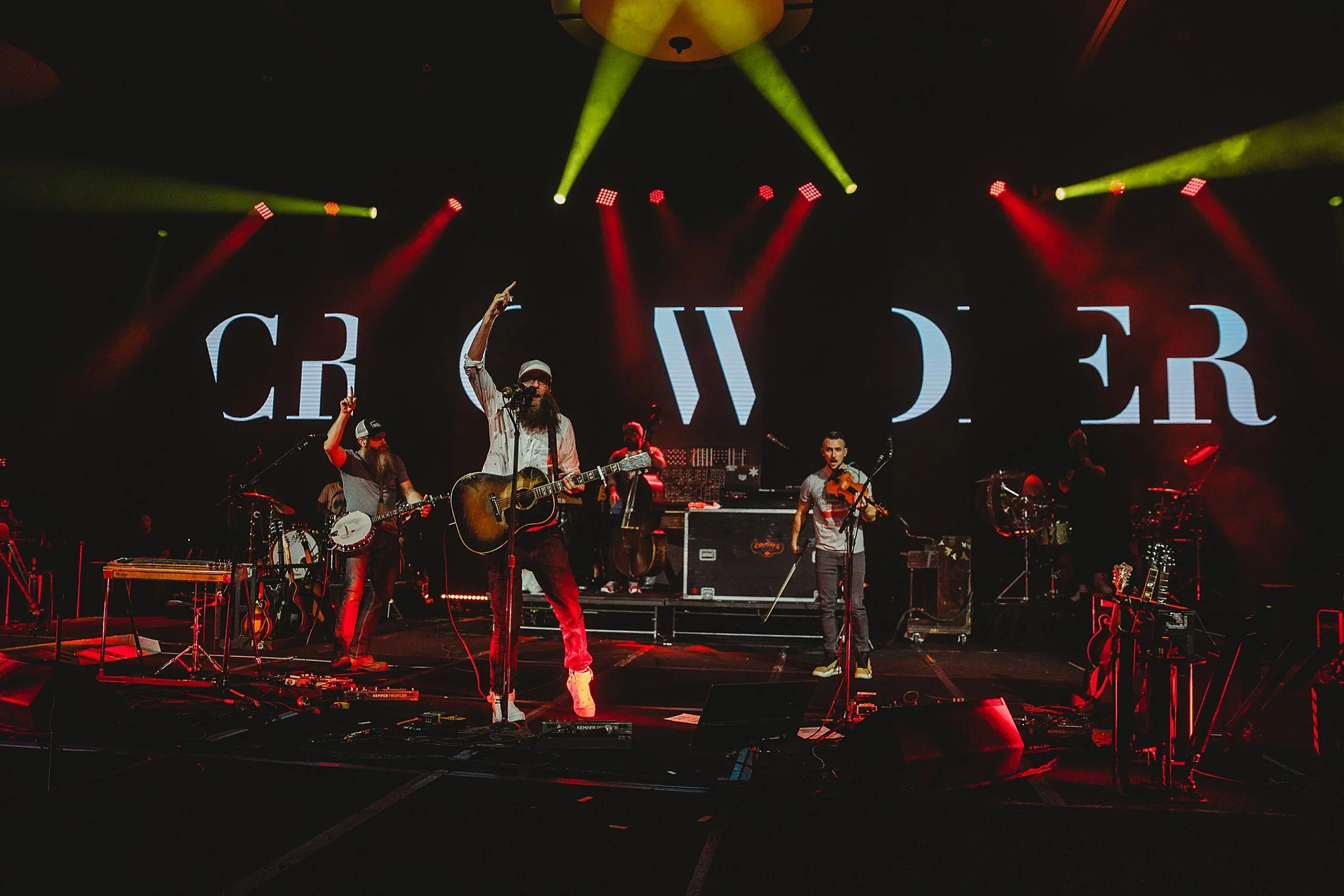 A band performing on stage with multiple musicians playing guitars, violin, and drums; stage is lit with yellow and red lights, with large screen in the background displaying the word "CREW" in stylized font.