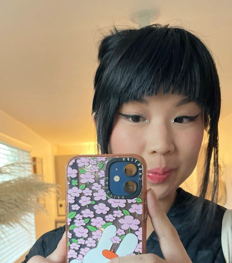 A woman with short black hair taking a selfie in a room with a smartphone that has a purple floral case. She has winged eyeliner and pink lipstick.