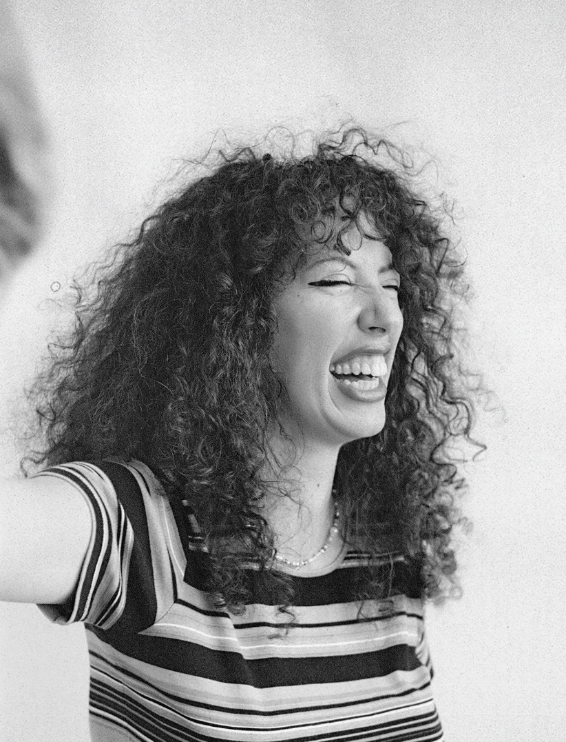 A woman with curly hair, smiling and laughing, wearing a striped shirt.