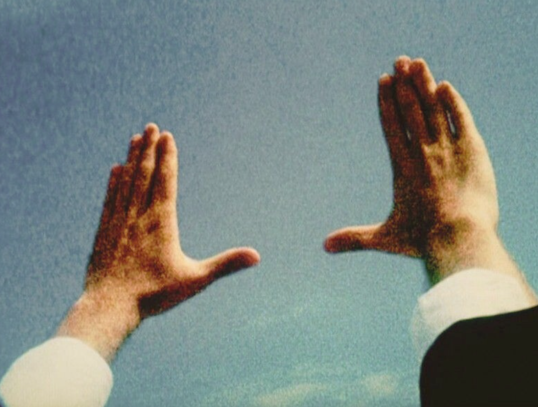 Two hands in formal attire positioned against a clear sky, with palms open and fingers spread apart.