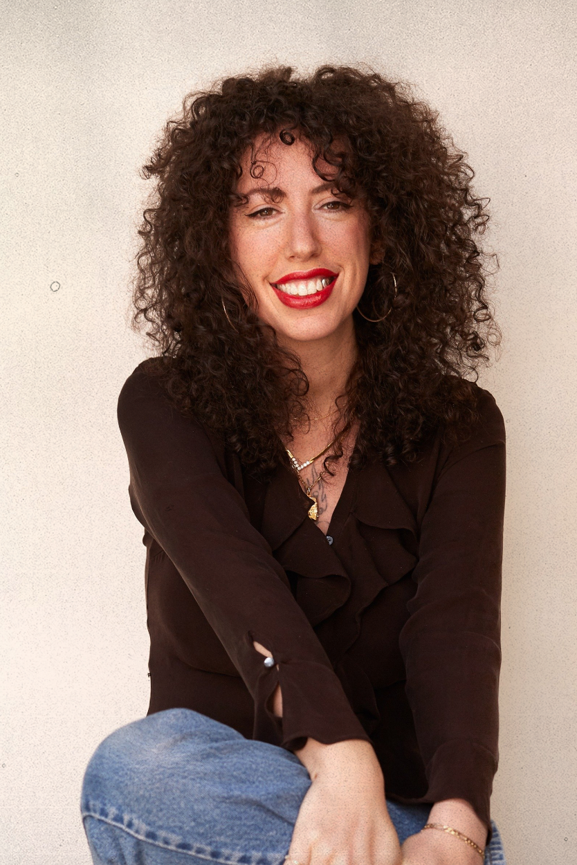A woman with curly dark hair, red lipstick, and hoop earrings, sitting and smiling at the camera against a plain wall, wearing a black blouse with ruffled details and blue jeans.