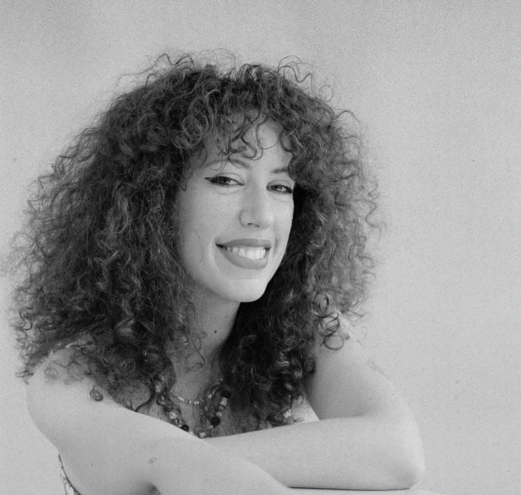 Black and white portrait of a young woman with curly hair smiling with arms crossed, sitting in front of a plain wall.