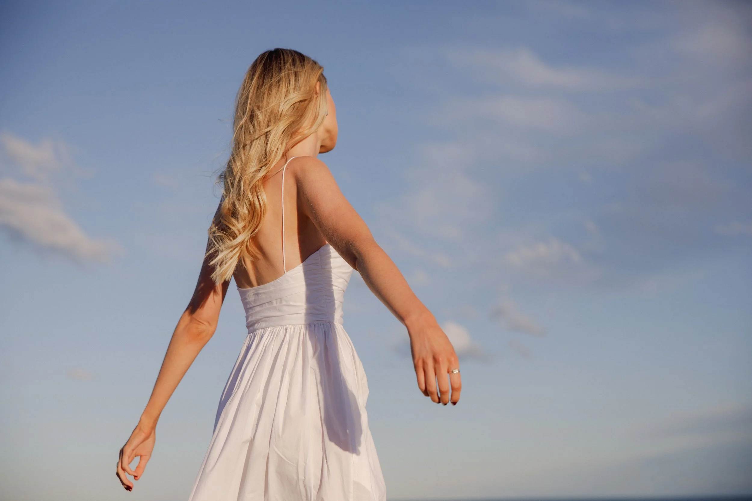 A woman in a white dress with long wavy blonde hair standing outdoors under a blue sky with scattered clouds, facing away from the camera.