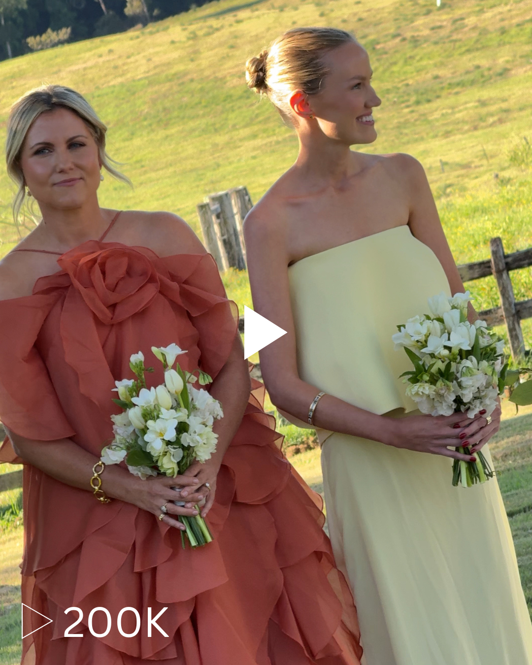 Two women in elegant dresses holding bouquets of white flowers, standing outdoors in a grassy field with a wooden fence in the background.