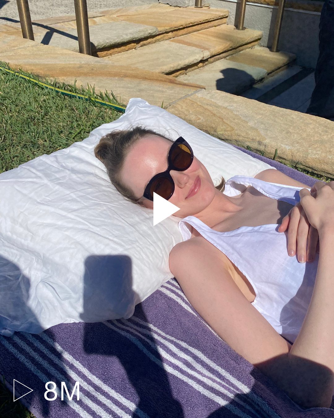 Woman lying on a pillow outdoors, wearing sunglasses and a white top, sunbathing on a purple towel.