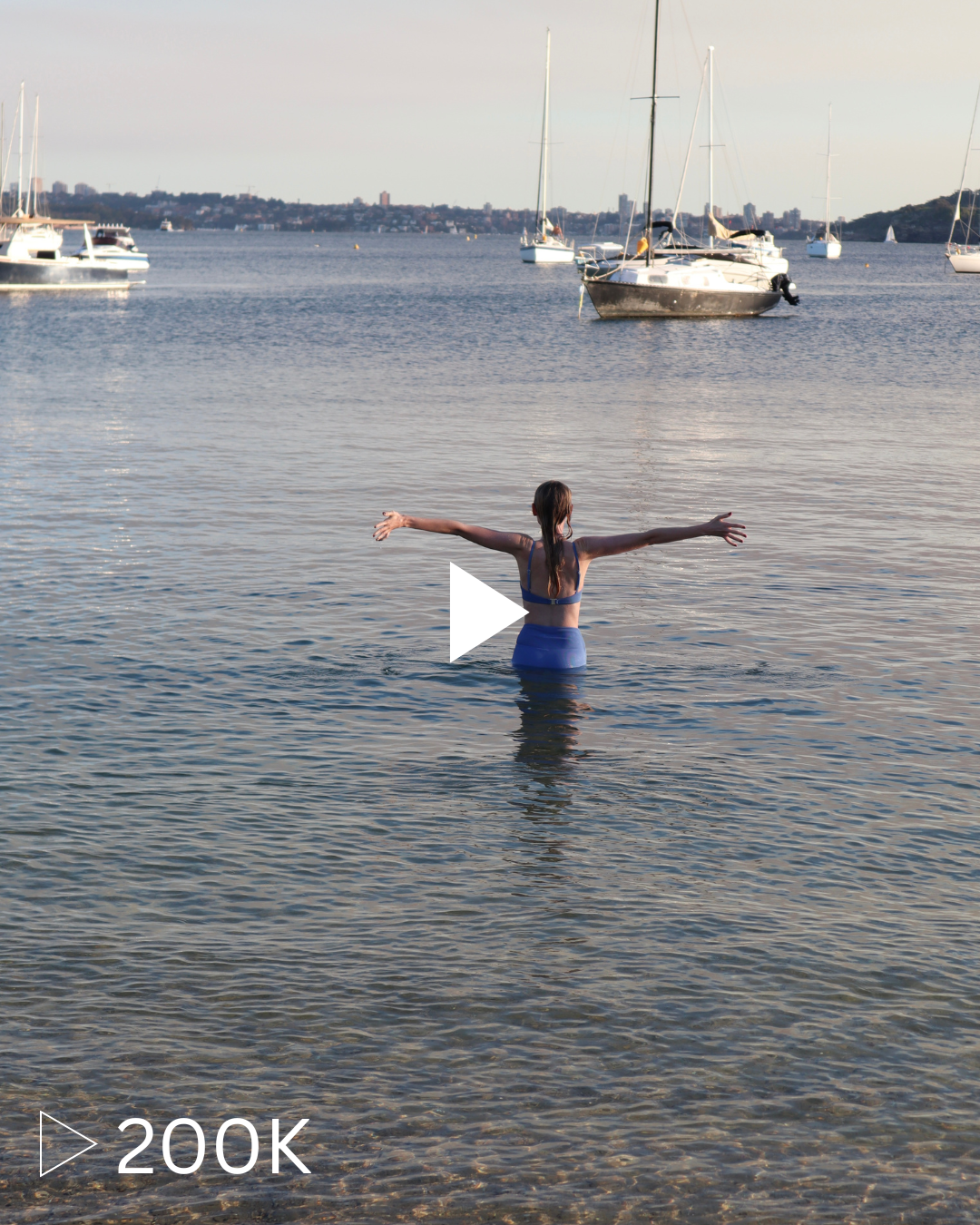 A woman in a blue swimsuit stands in the water with her arms outstretched, facing a marina with sailboats anchored in the distance.