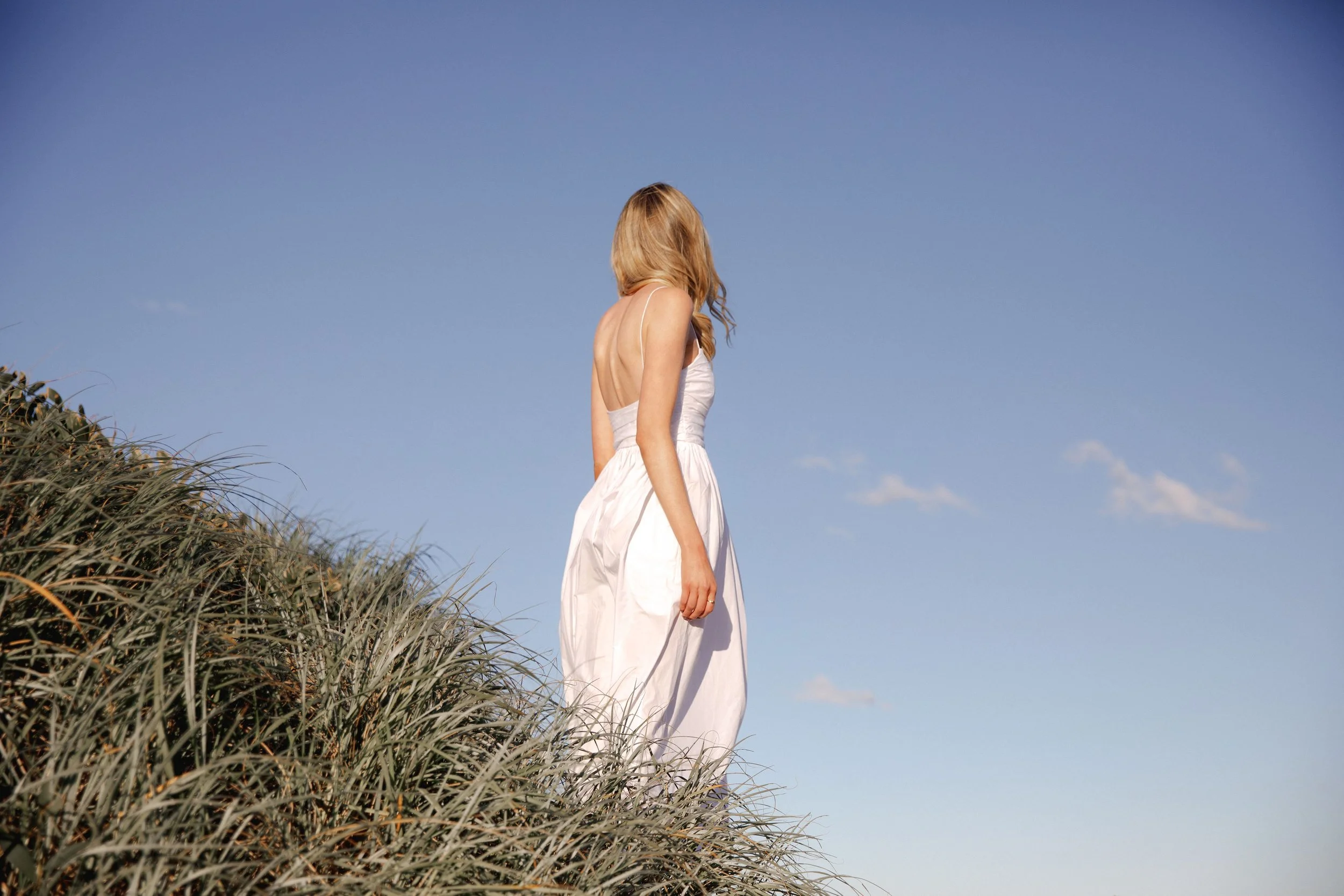 Lily Schubert, Sydney keynote speaker and content creator, wearing a white dress standing among tall grasses outdoors, facing away from the camera, under a clear blue sky.