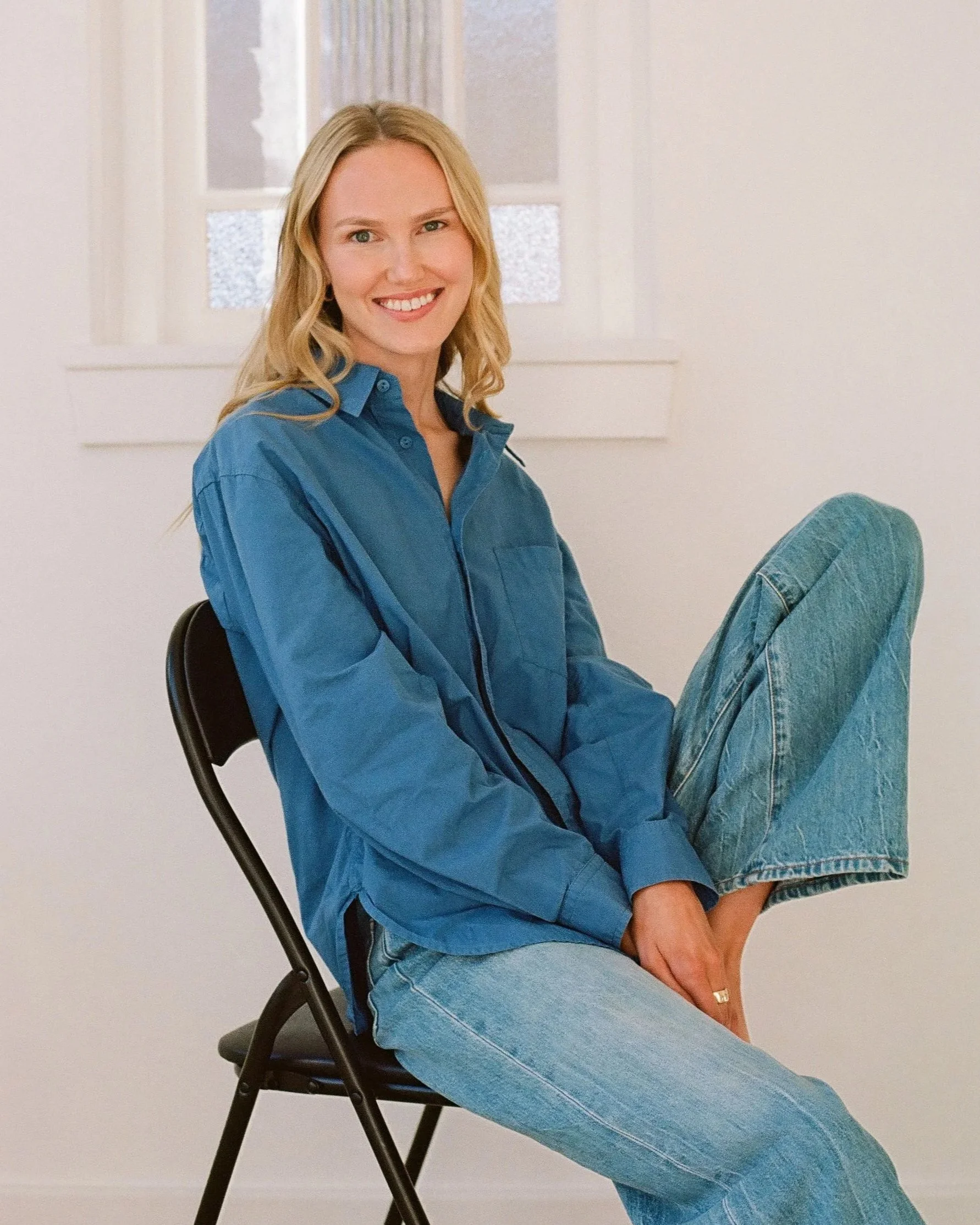 Lily Schubert, Sydney keynote speaker and content creator, smiling, sitting on a black chair, wearing a blue jacket and blue jeans, with a white wall and window in the background.