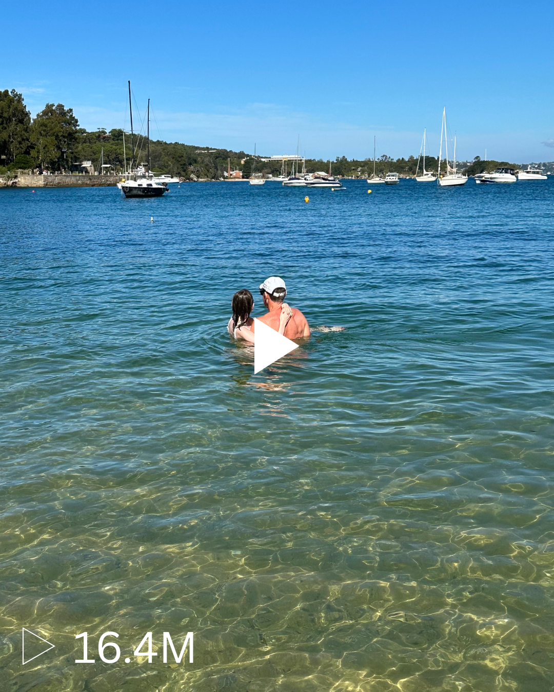 A man and a young girl are swimming and embracing in clear, shallow water near a dock, with boats anchored in the distance on a sunny day.