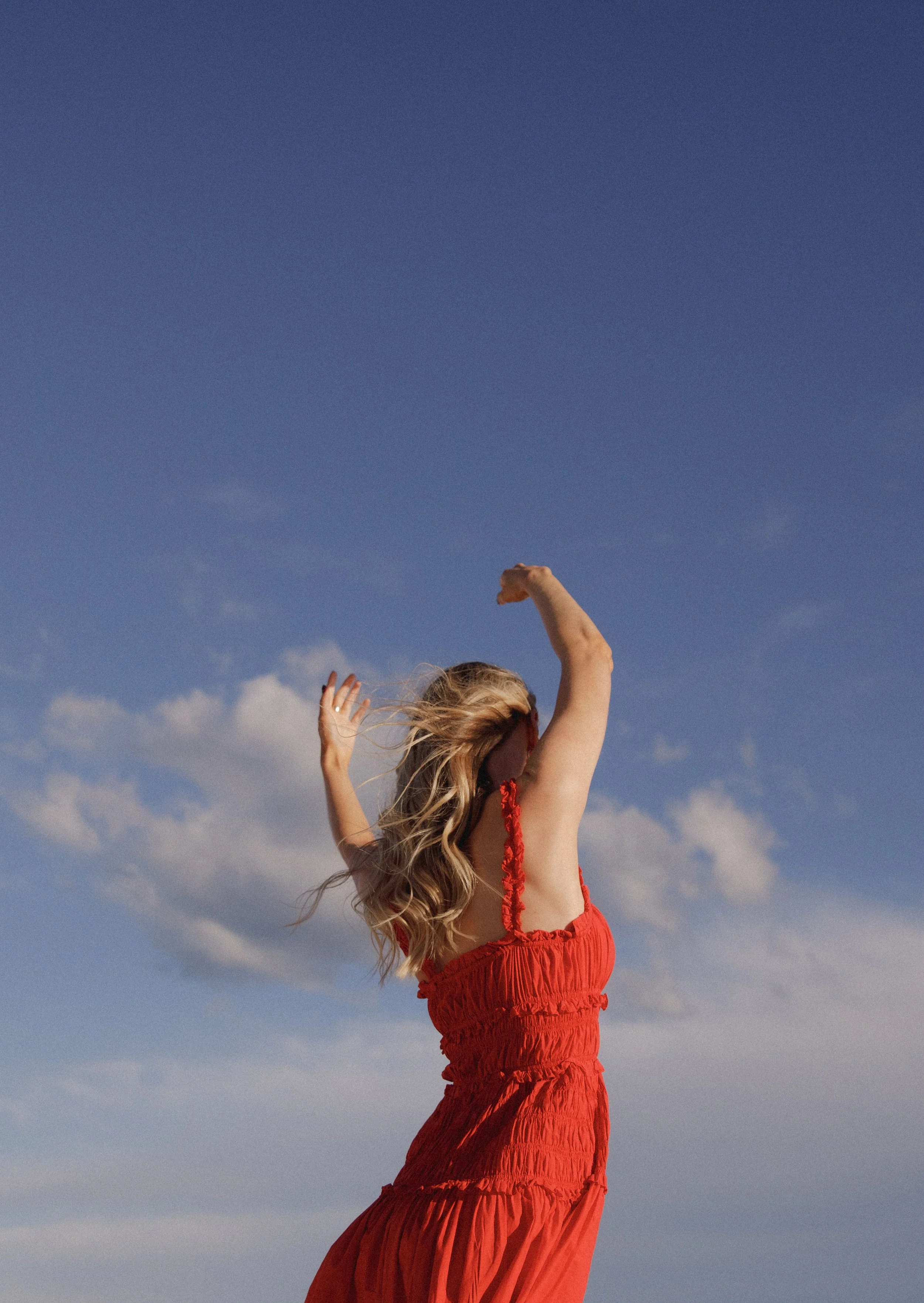 Lily Schubert, Sydney keynote speaker and content creator with long blonde hair wearing a red dress with ruffles raises her arms in the air against a background of blue sky with scattered clouds.