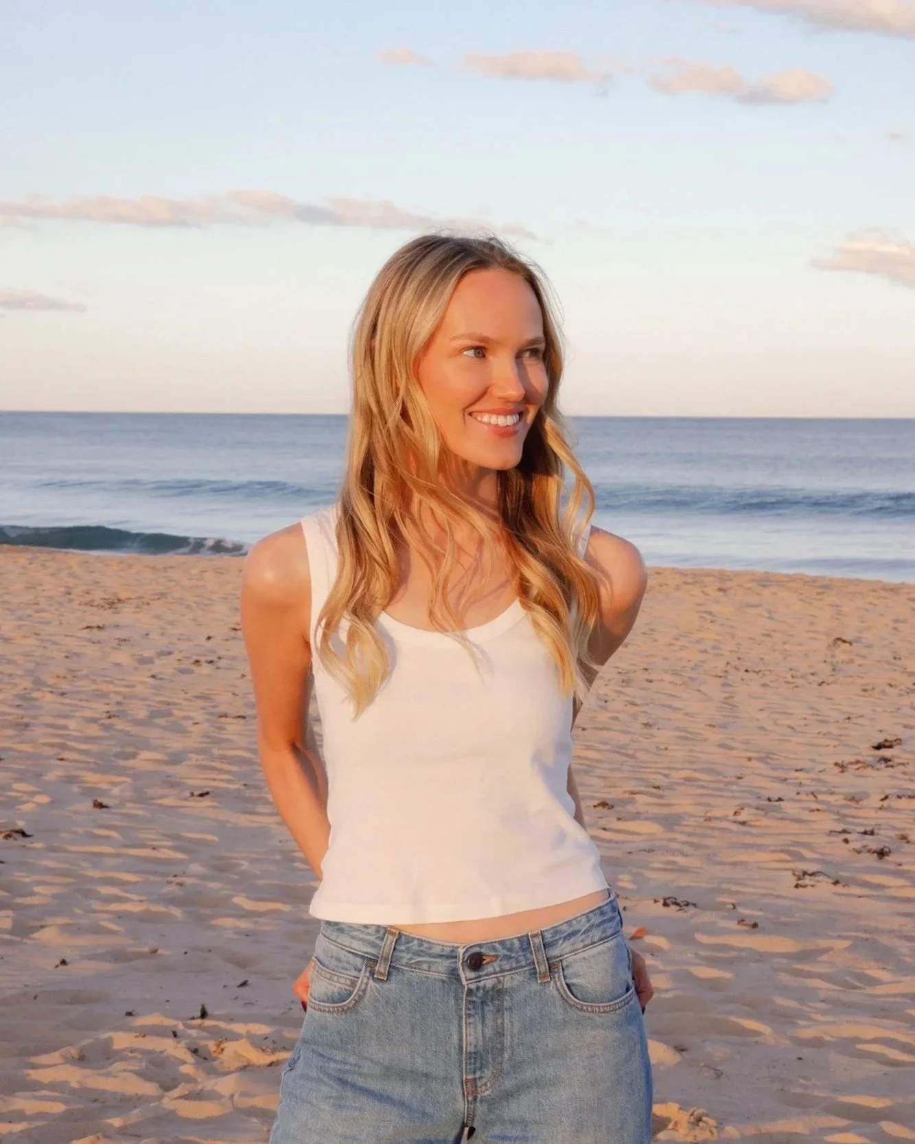 Lily Schubert standing on a sandy beach near the ocean with waves and a partly cloudy sky in the background.