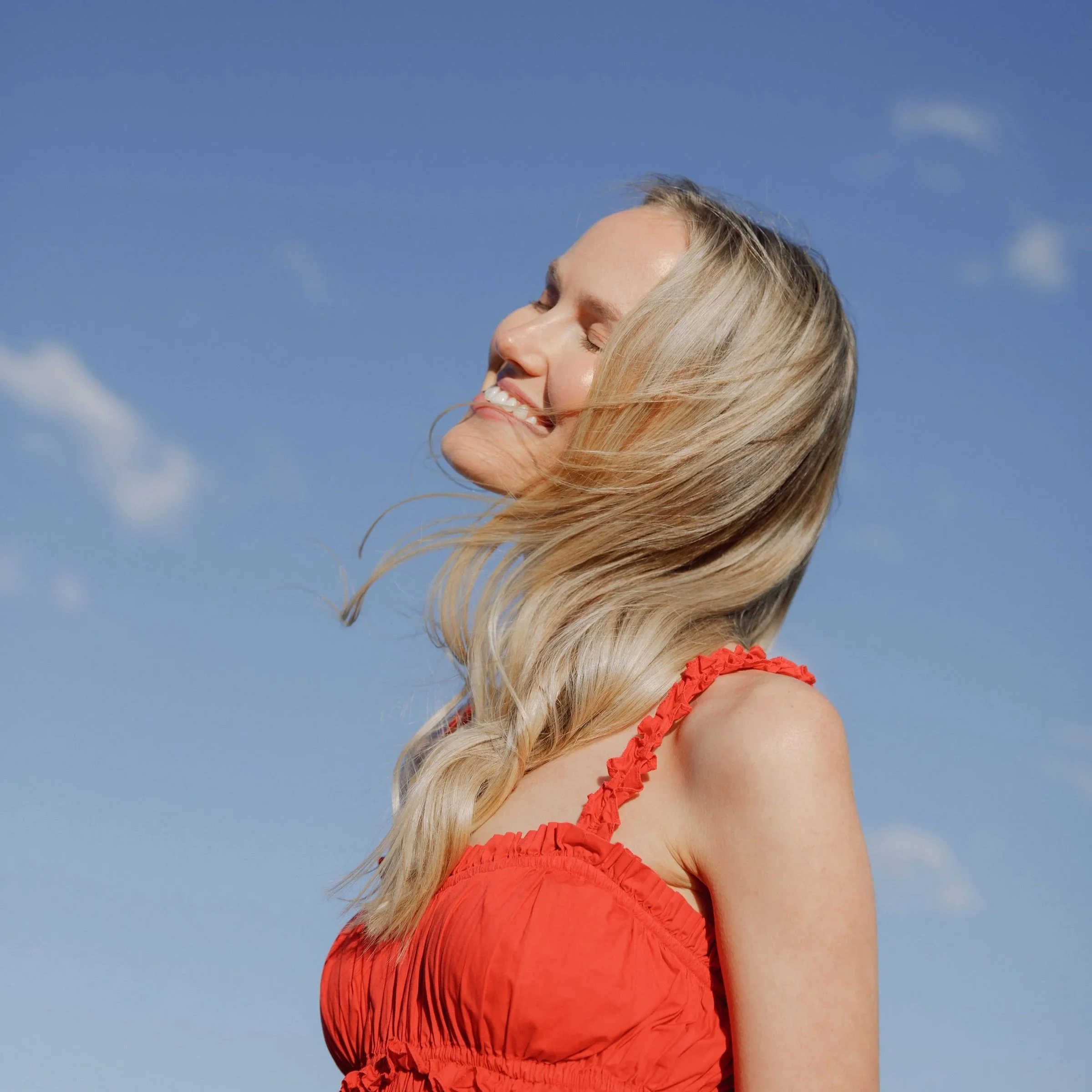 Lily Schubert, keynote speaker and content creator,  smiling with her eyes closed, wearing a ruffled orange dress, against a background of a clear blue sky with a few clouds.