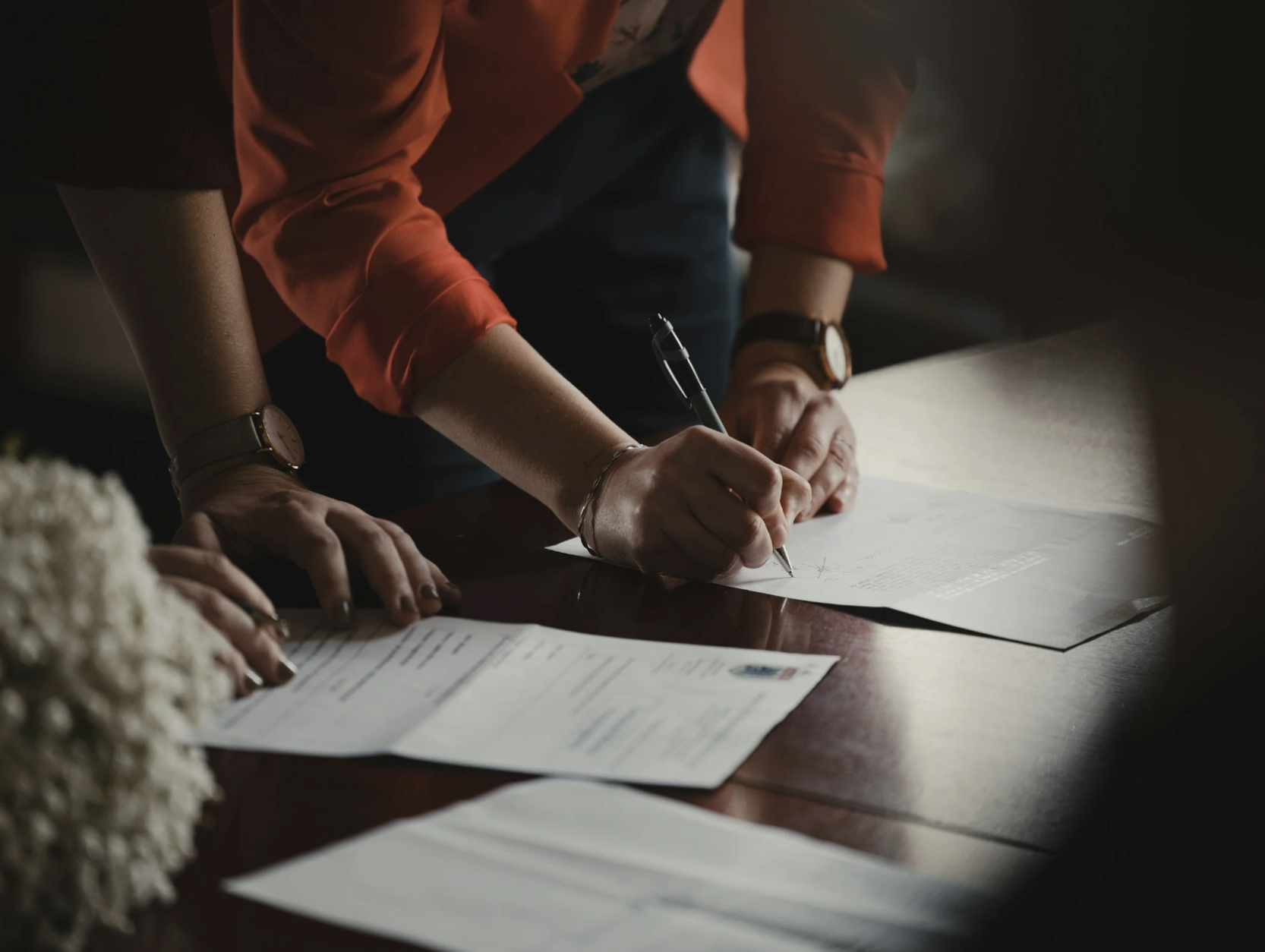 Person in an orange shirt signing a document on a dark wooden table with papers and a flower arrangement nearby.