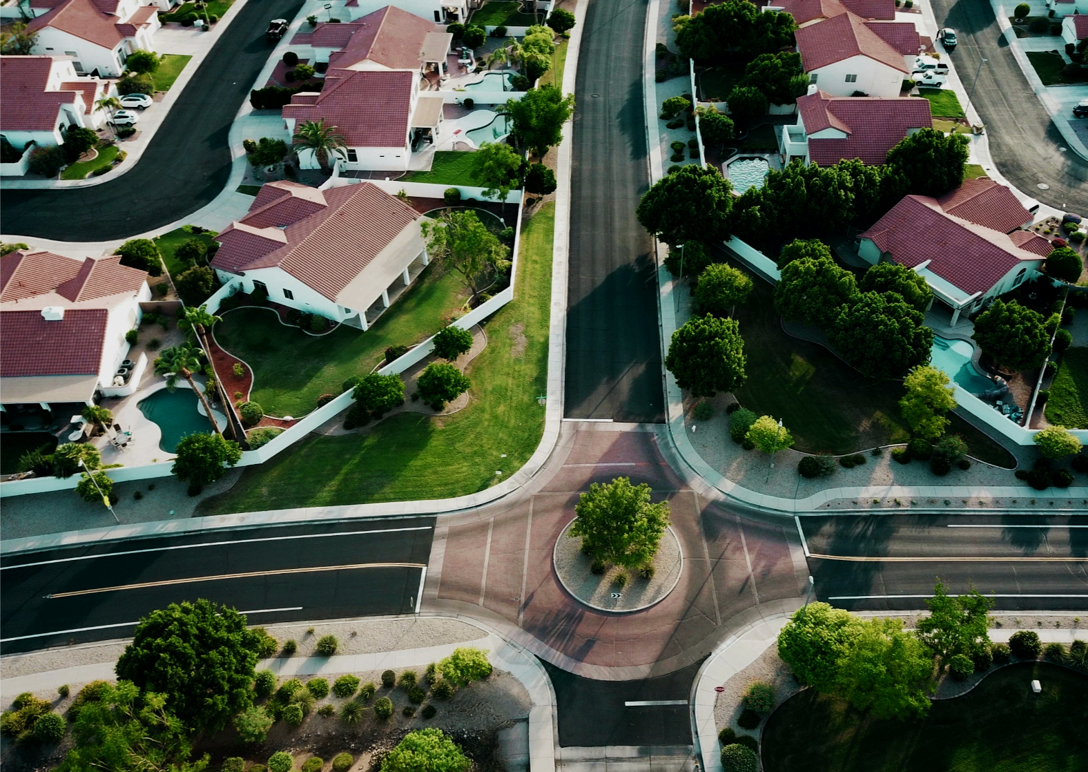 An aerial view of a residential neighborhood with houses, trees, and asphalt roads, featuring a landscaped roundabout with a tree at the center.