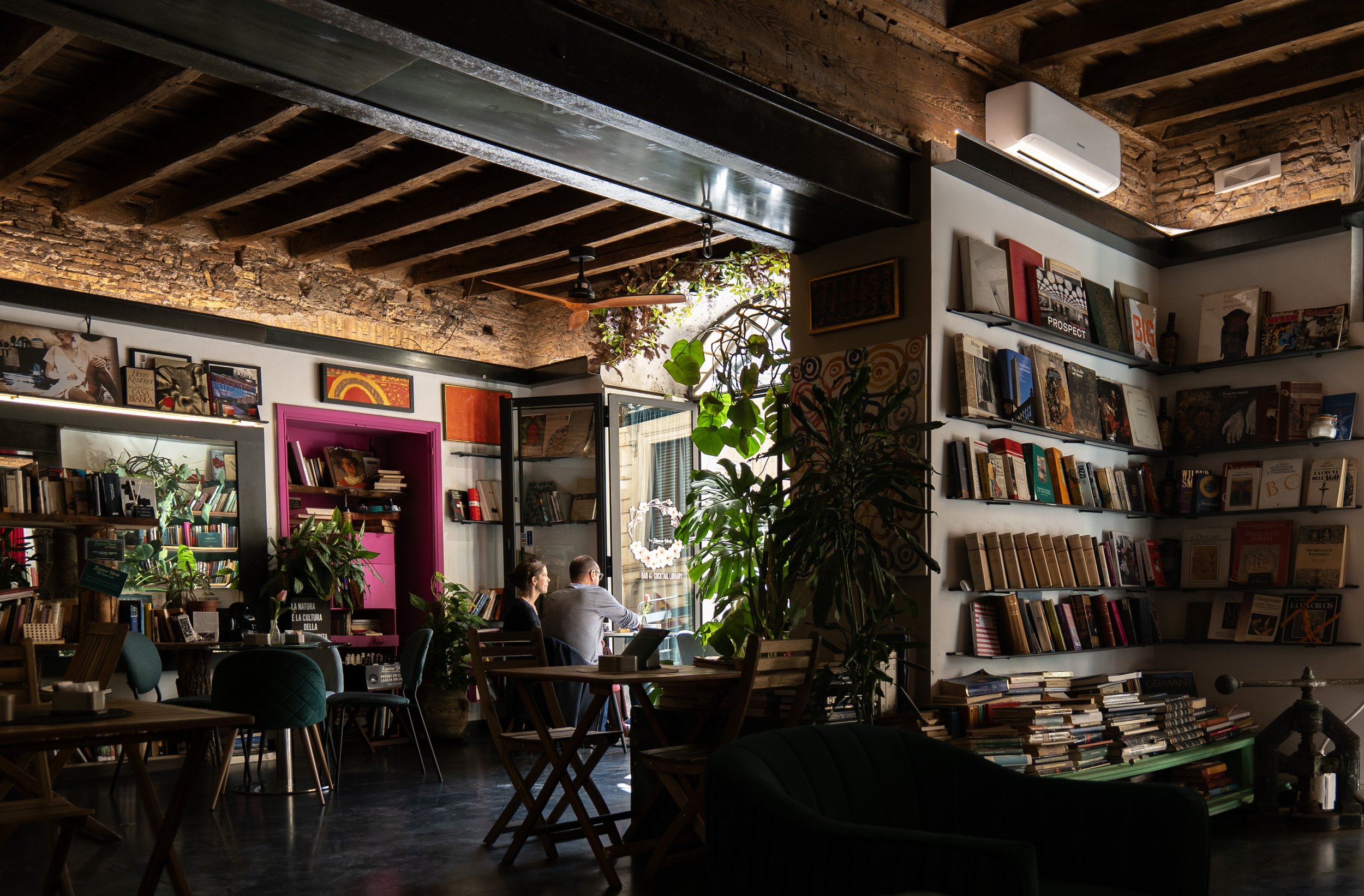 Interno di una libreria o caffè con scaffali pieni di libri, divani e tavoli, alcune persone sedute a uno di questi tavoli che lavorano o leggono, piante verdi e pareti in mattoni.