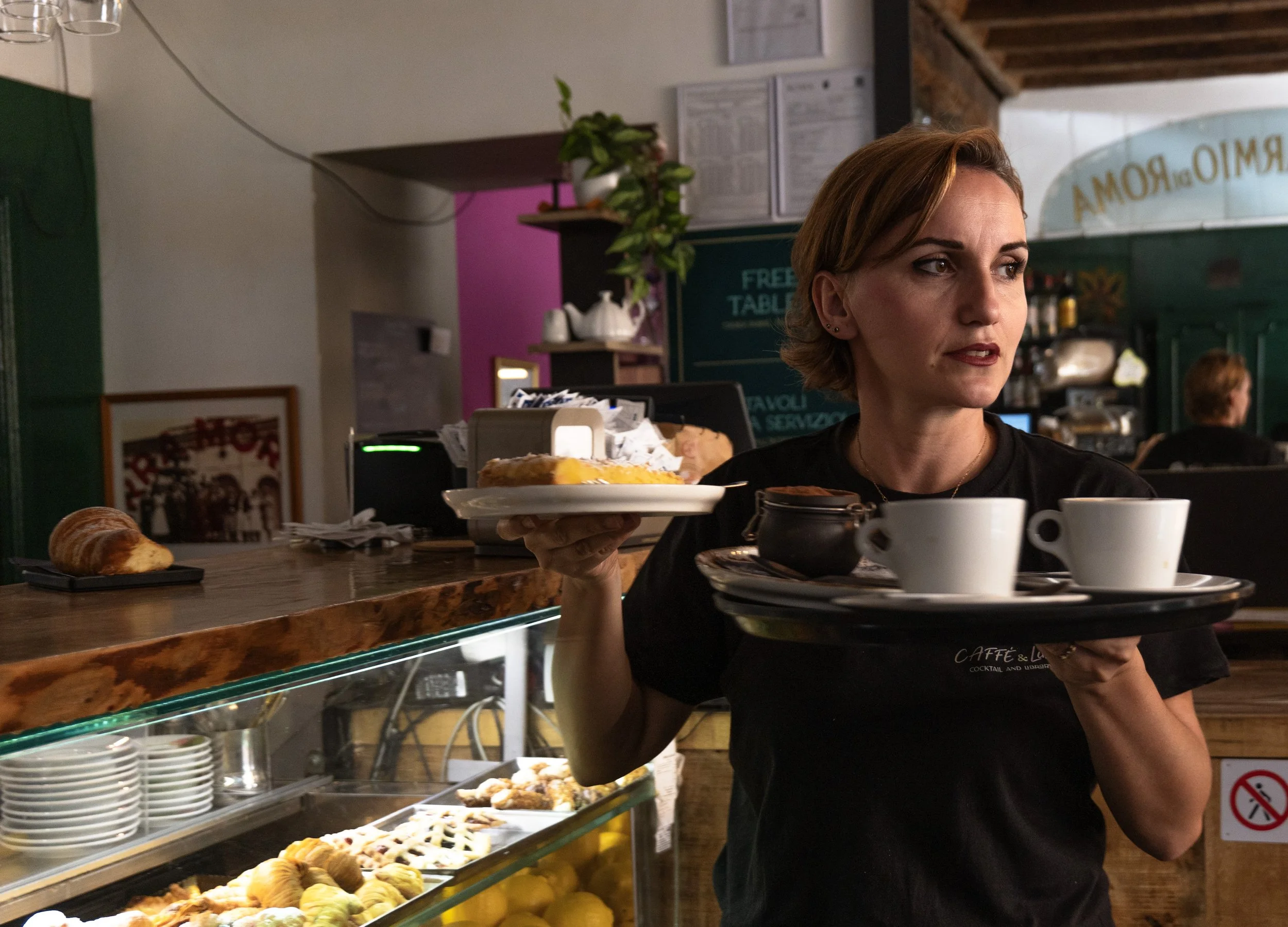 Una donna in un bar o caffetteria, che tiene due vassoi con tazze e piatti di cibo e bevande, tra cui un dolce e una cioccolata.