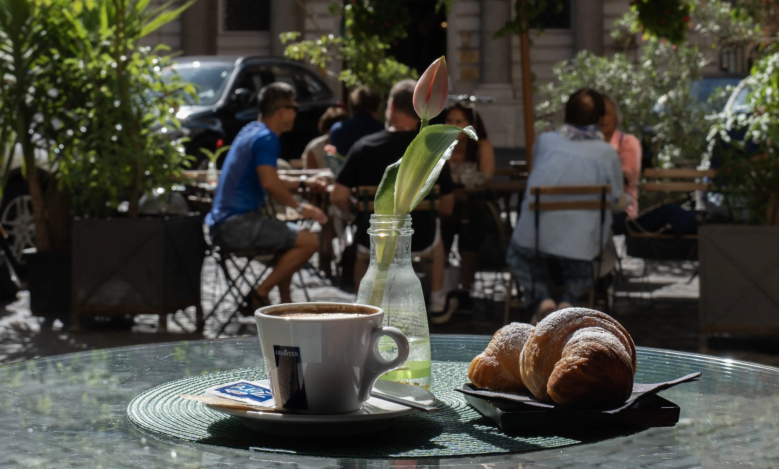 Tazza di caffè, croissant e una pianta in un vaso su un tavolo di un bar all'aperto, con persone sedute e chiacchierando sullo sfondo.