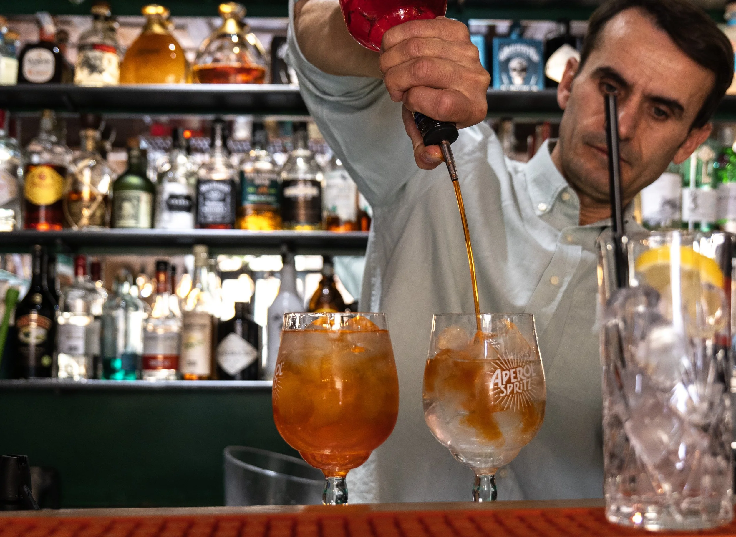 Un bartender versa un liquido in un bicchiere con ghiaccio e una fetta di limone, mentre altri due bicchieri con bevande arancioni sono sul bancone di un bar.
