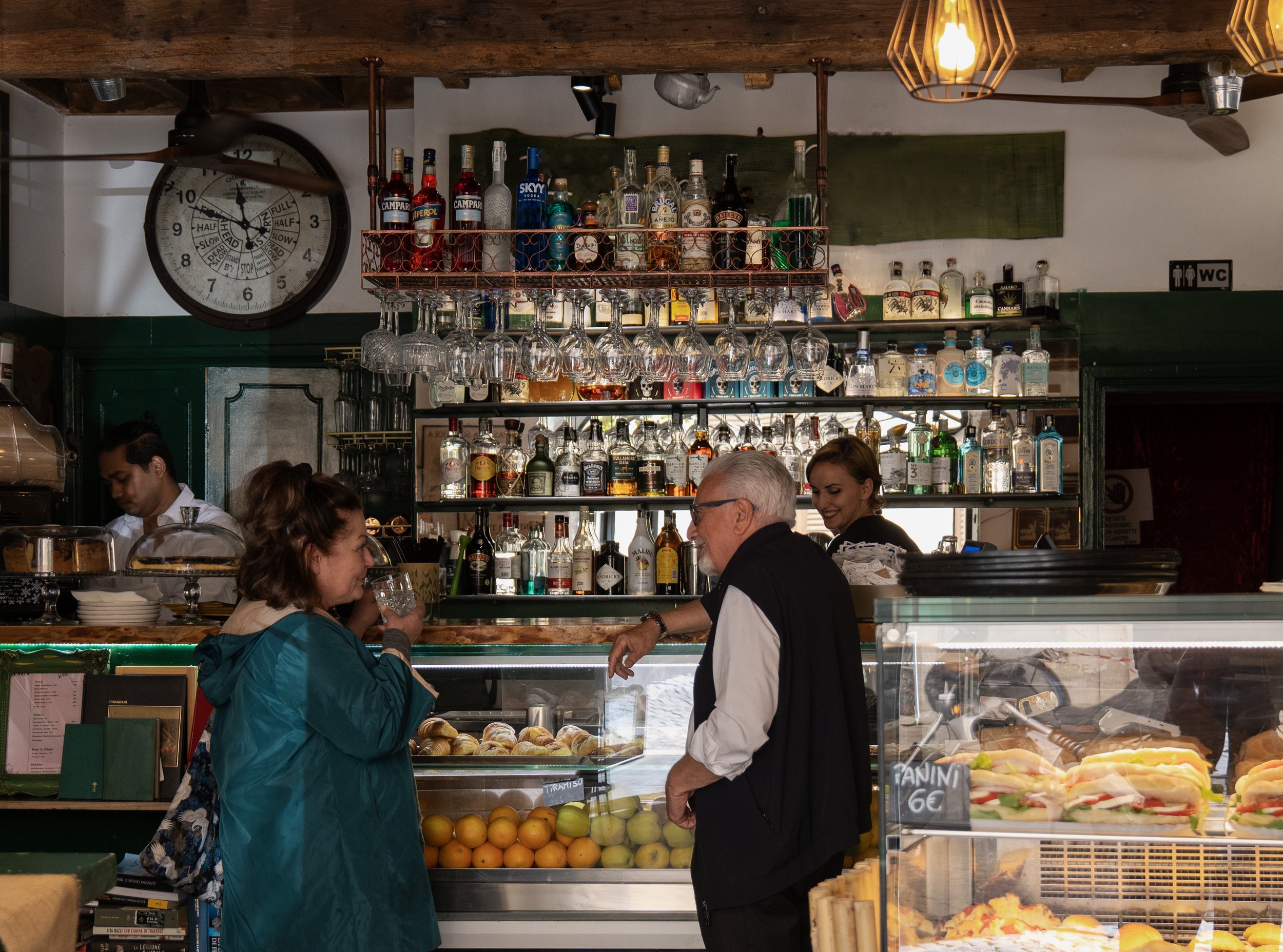 Interno di un bar con persone che chiacchierano, scaffali di bottiglie di alcolici sullo sfondo, frutta e panini nel caso davanti, e un grande orologio sulla parete a sinistra.