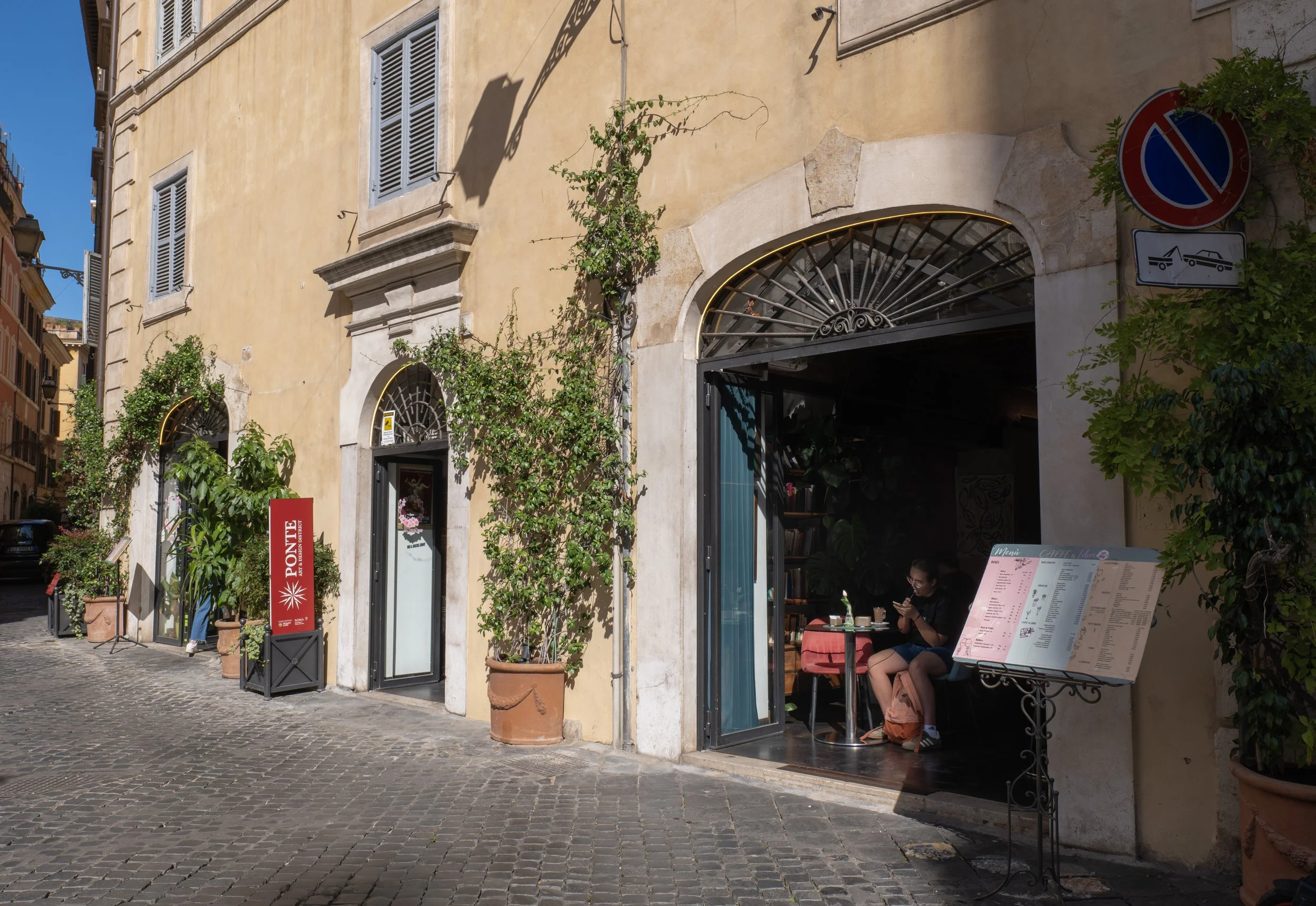 Strada in pietra con edifici storici, un caffè con clienti seduti e menu all'esterno, e segnali di divieto di sosta.
