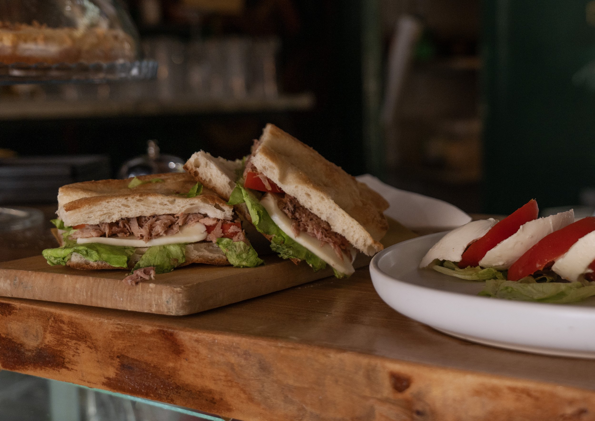 Panino con carne, lattuga, pomodoro e maionese su tagliere di legno accanto a una caprese con mozzarella, pomodoro e lattuga in una ciotola bianca.