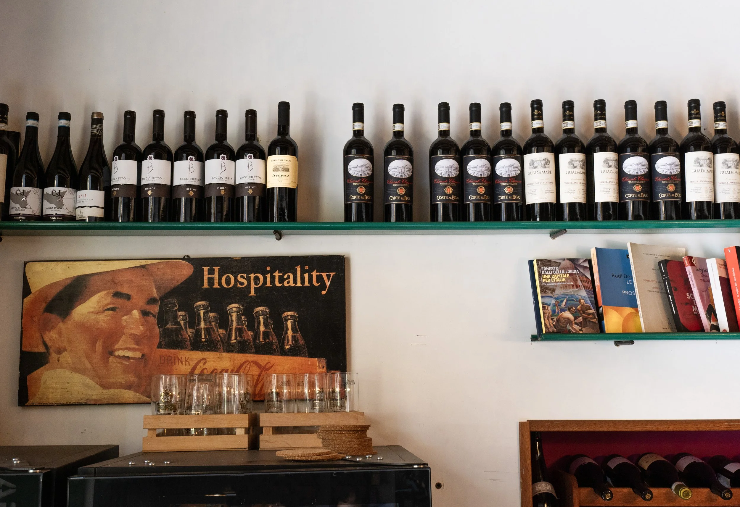 Vasche di acqua, un cartello vintage con l'immagine di una donna sorridente e la scritta 'Hospitality', una mensola con libri, e scaffali con bottiglie di vino.