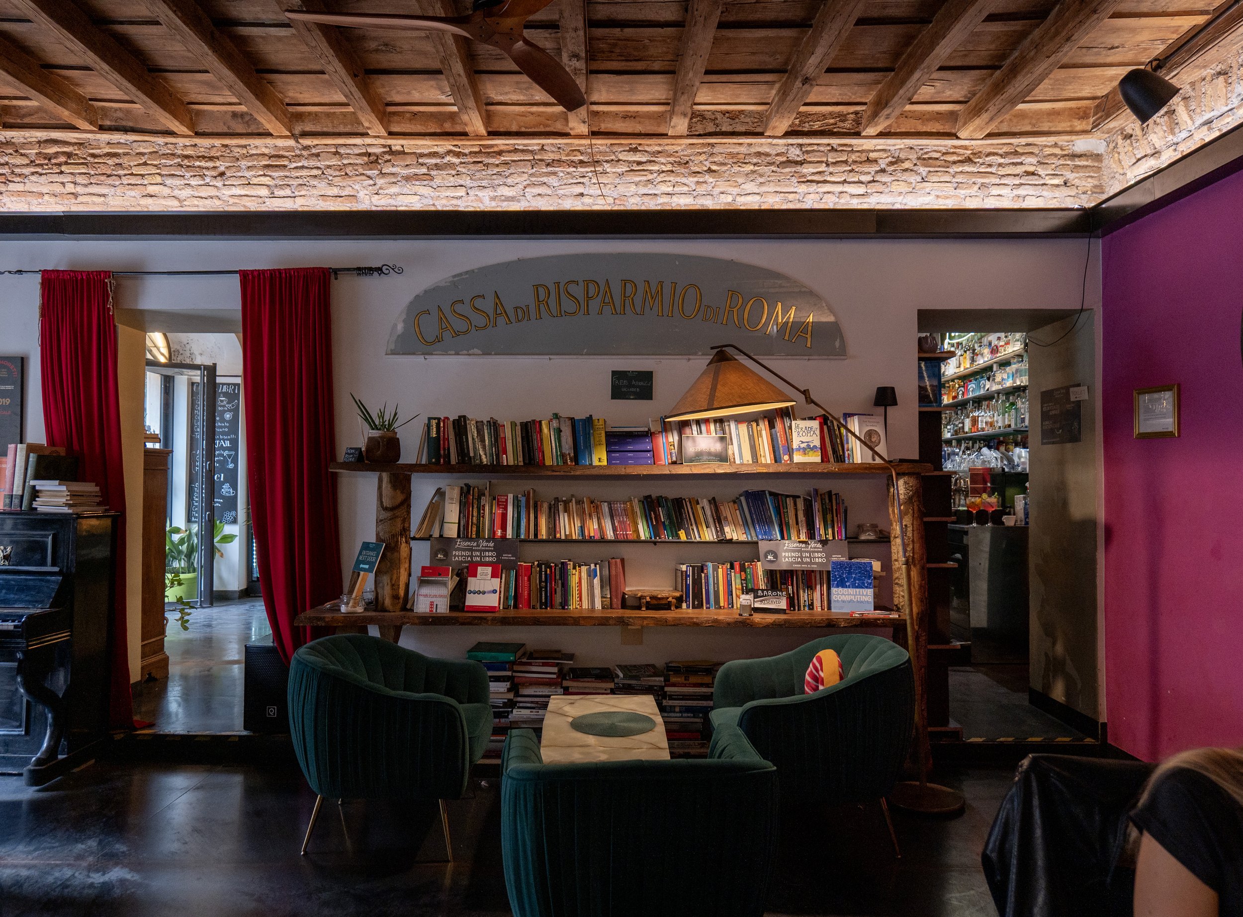 Interno di una libreria con scaffali pieni di libri, poltrone verdi intorno a un tavolino, parete rosa e parete di mattoni a vista, con scritto 'Cassa di Risparmio di Roma'.