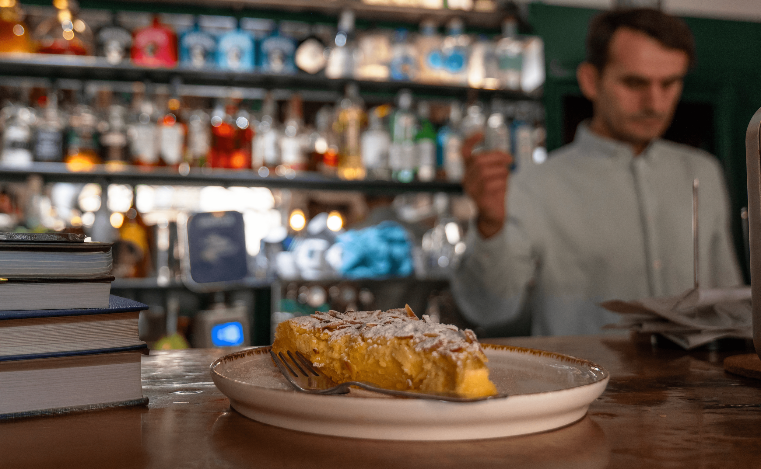Torta di crema alla vaniglia con zucchero a velo su un piatto, con forchetta, davanti a un bar con bottiglie di alcolici sullo sfondo e un uomo dietro al banco.