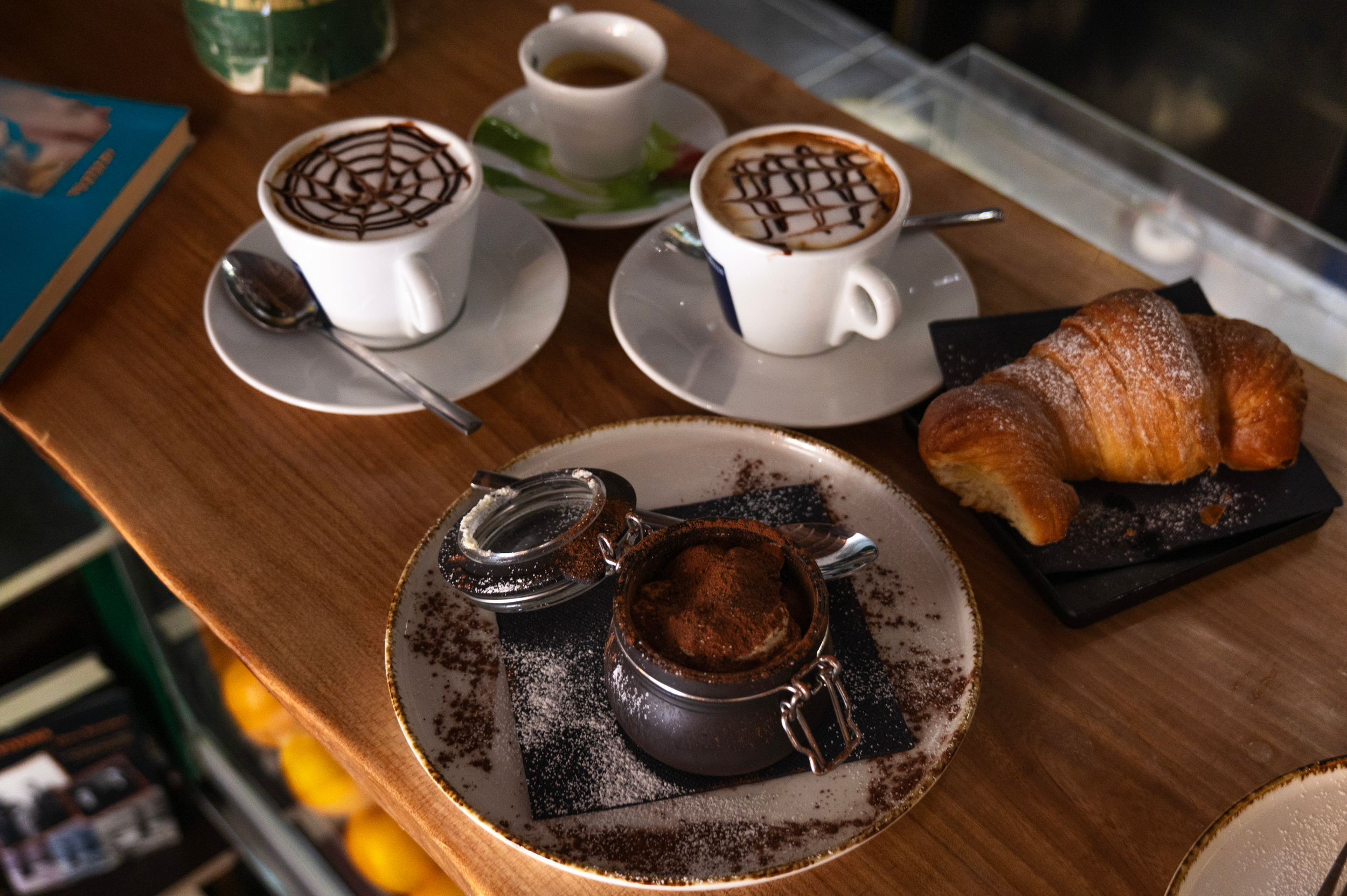 Tazza di cappuccino con decorazione di cioccolato, altra tazza di cappuccino con decorazione, croissant, dolce al cioccolato in una scatola di metallo con zucchero a velo, tazza di caffè, libro blu su un tavolo di legno.