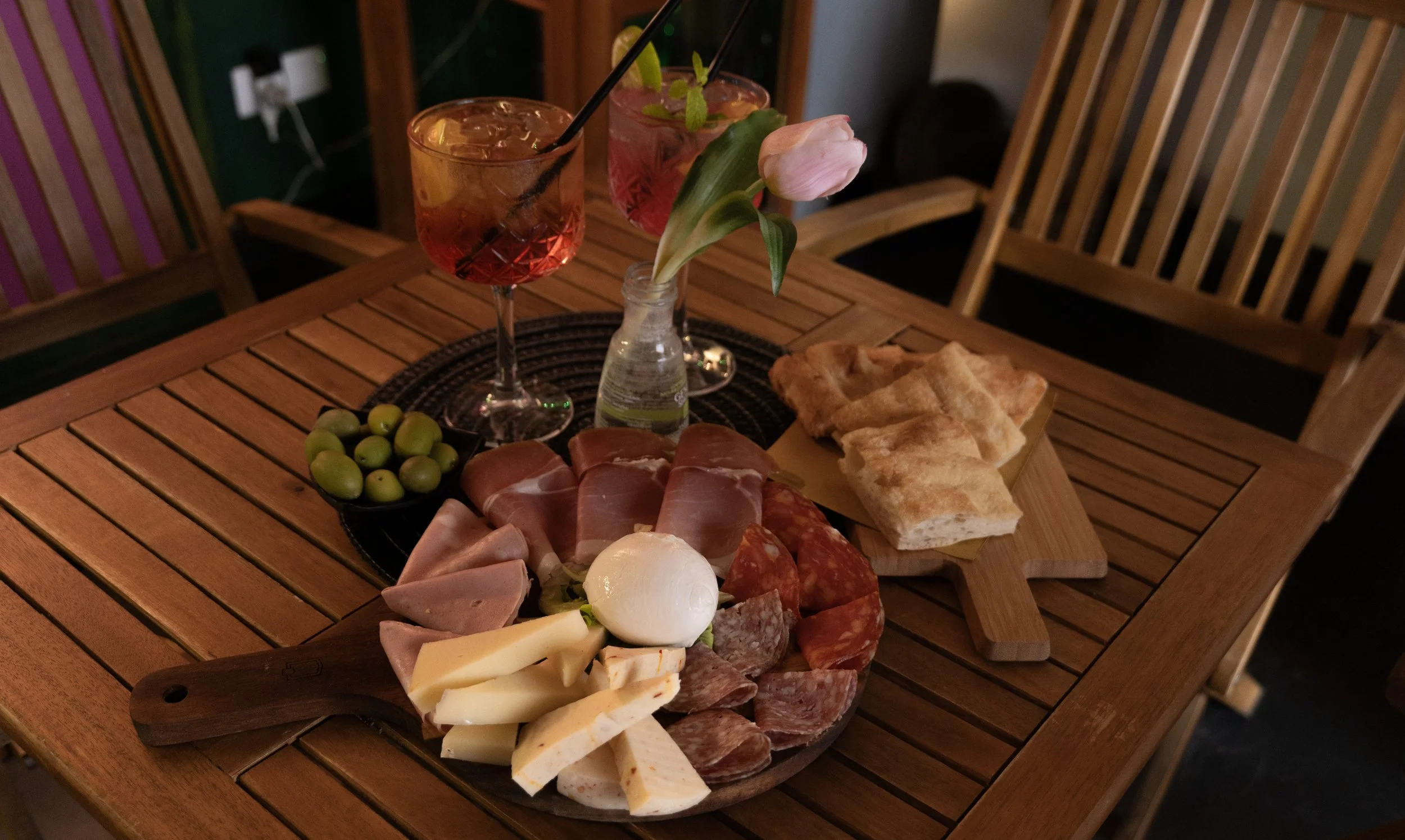 Tagliere di salumi e formaggi con pane, olive verdi, due cocktail rosati con limone e menta, fiore di tulipano rosa in vaso, in una stanza con sedie di legno.