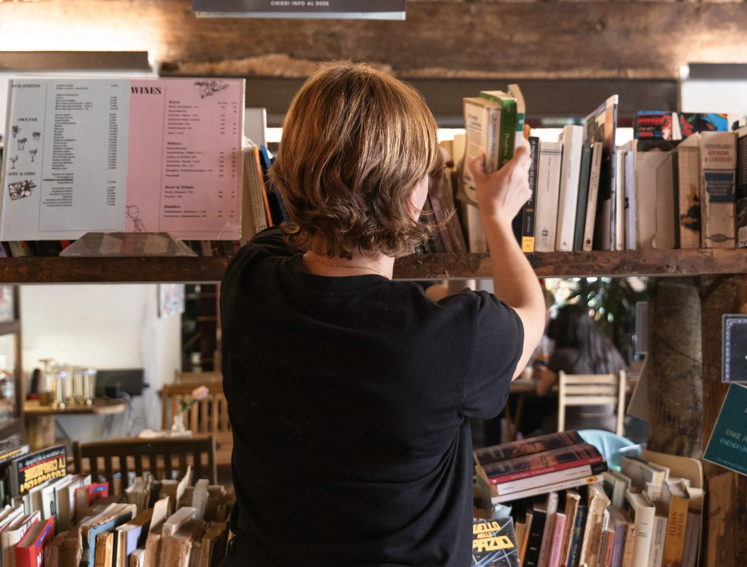 Una donna con capelli castani corti che guarda e prende un libro dagli scaffali di una libreria.